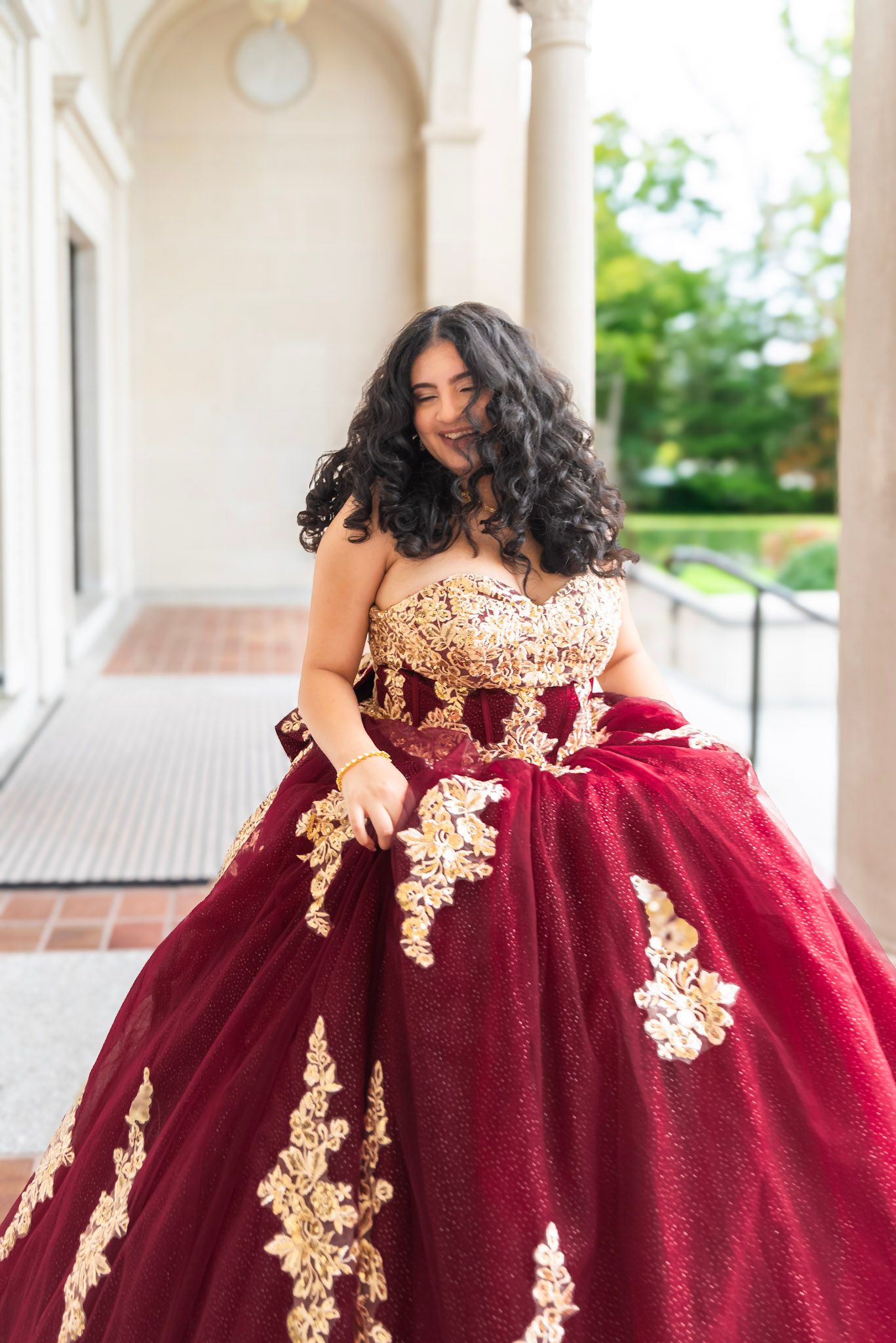 A burgundy and gold quinceañera dress with intricate lace details photographed on an elegant porch.