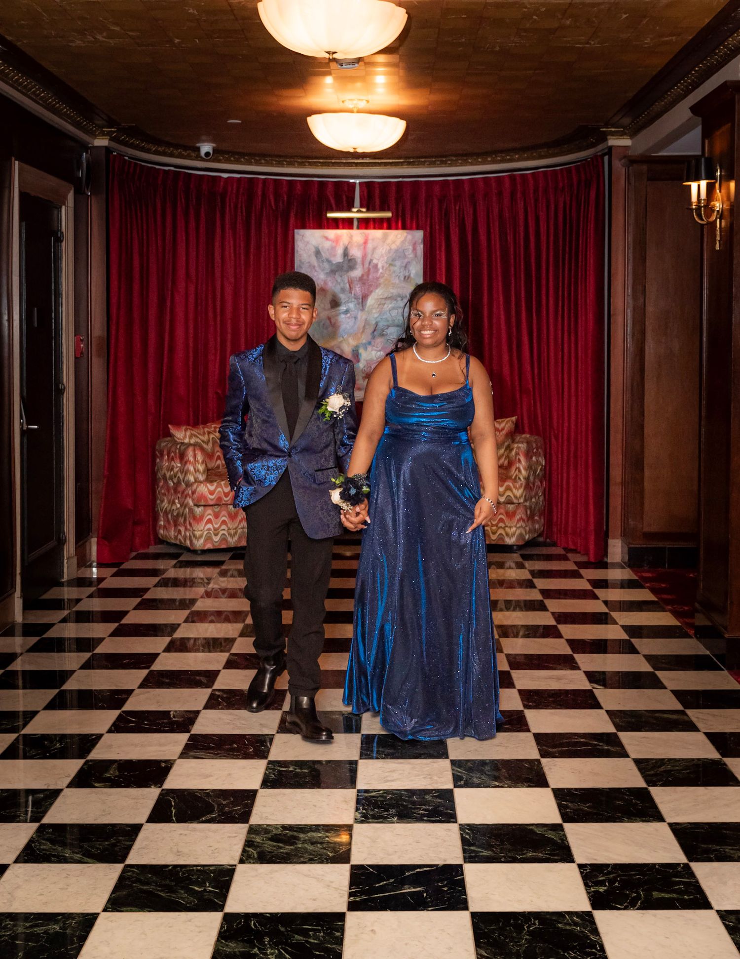 Couple walking on elegant black and white checkered floor with red curtains and chandelier lighting in formal venue.