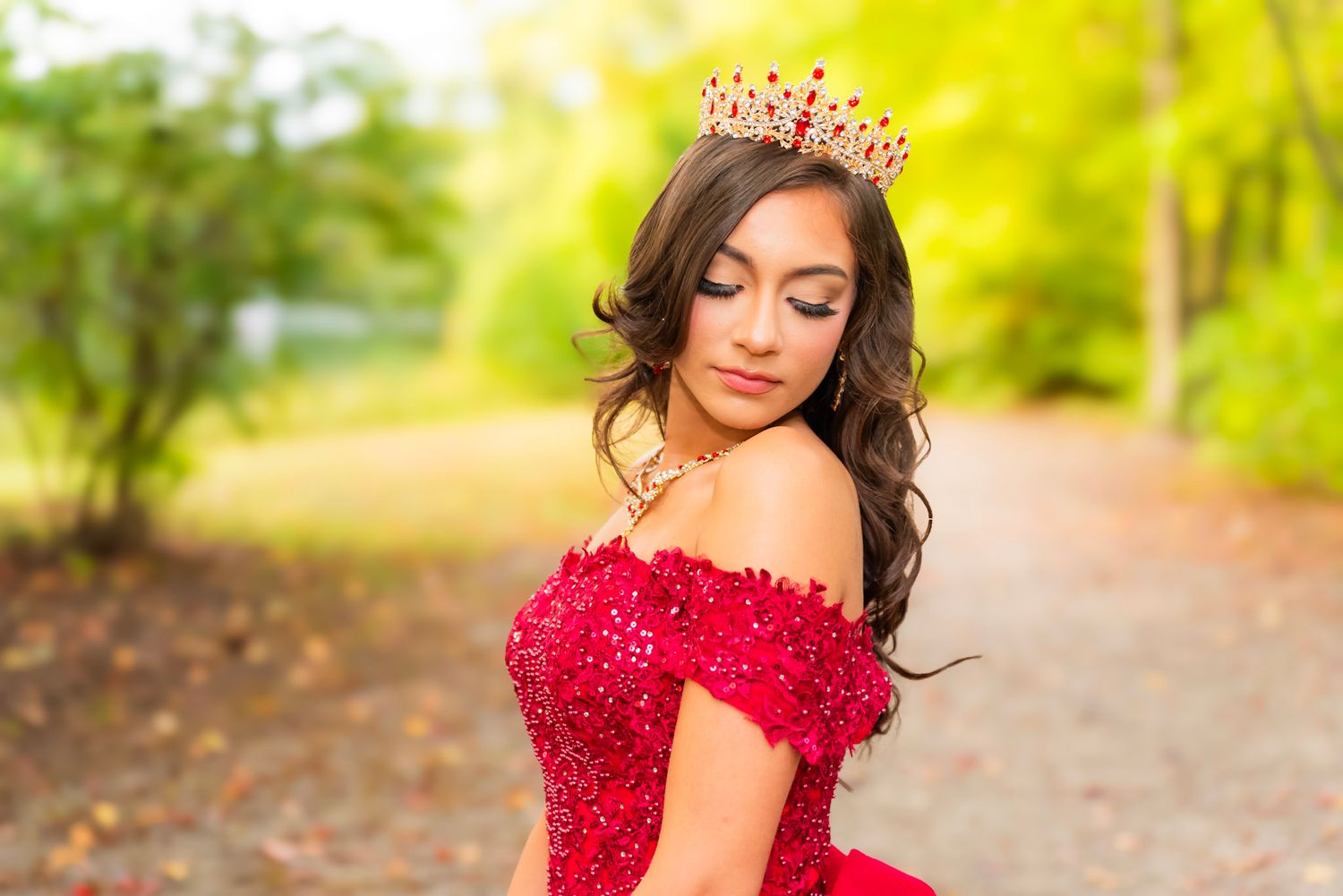 An elegant red off-shoulder ballgown with beaded details photographed in a sunny outdoor garden setting.
