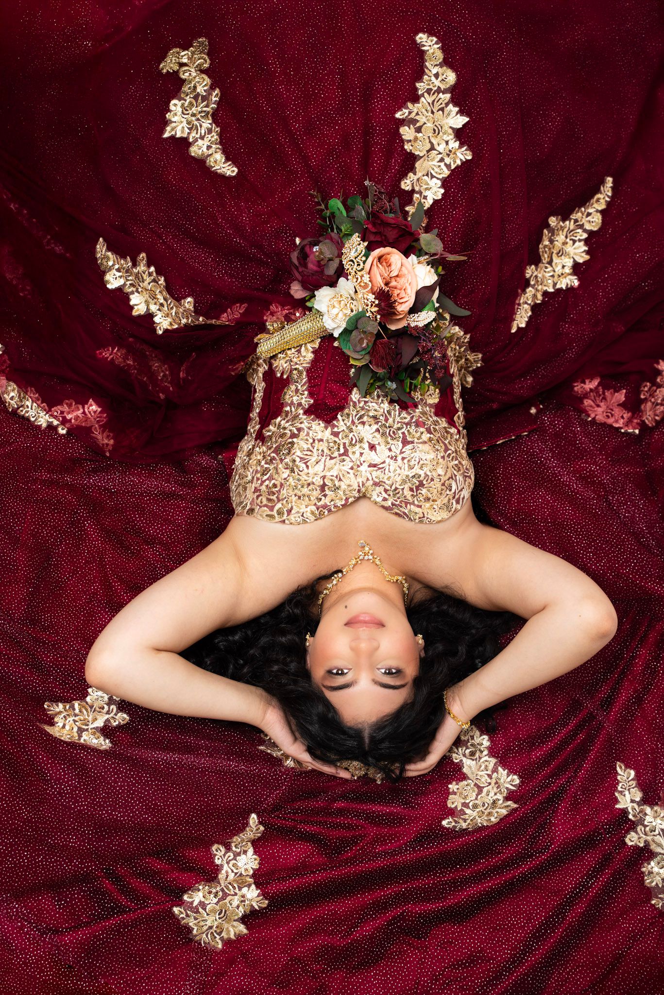 A person lying on burgundy velvet fabric with white flowers scattered around in an artistic portrait composition.