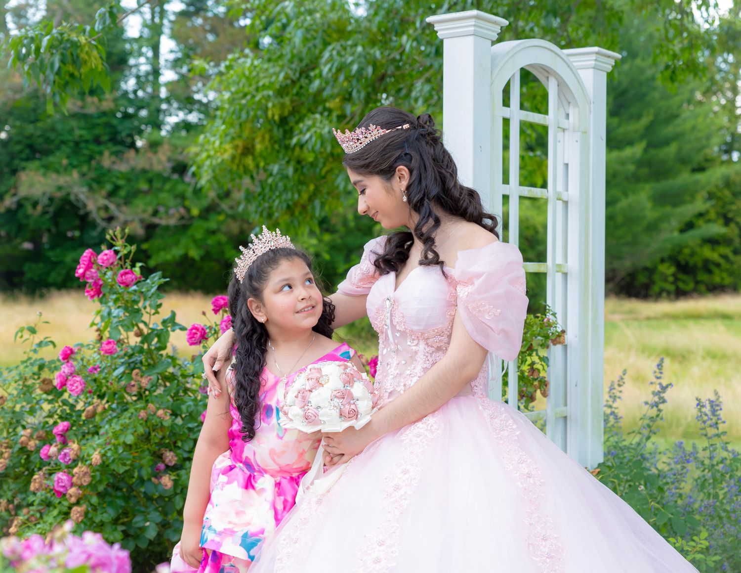 Elegant outdoor Quinceañera photo setting with white pergola and pink climbing roses in garden. Sibling girls looking at each other smiling