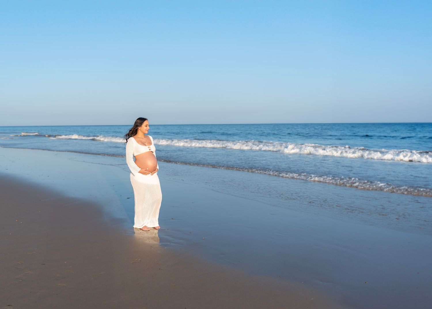 Maternity Photoshoot featuring Serene beach scene with waves crashing on shore under blue sky.