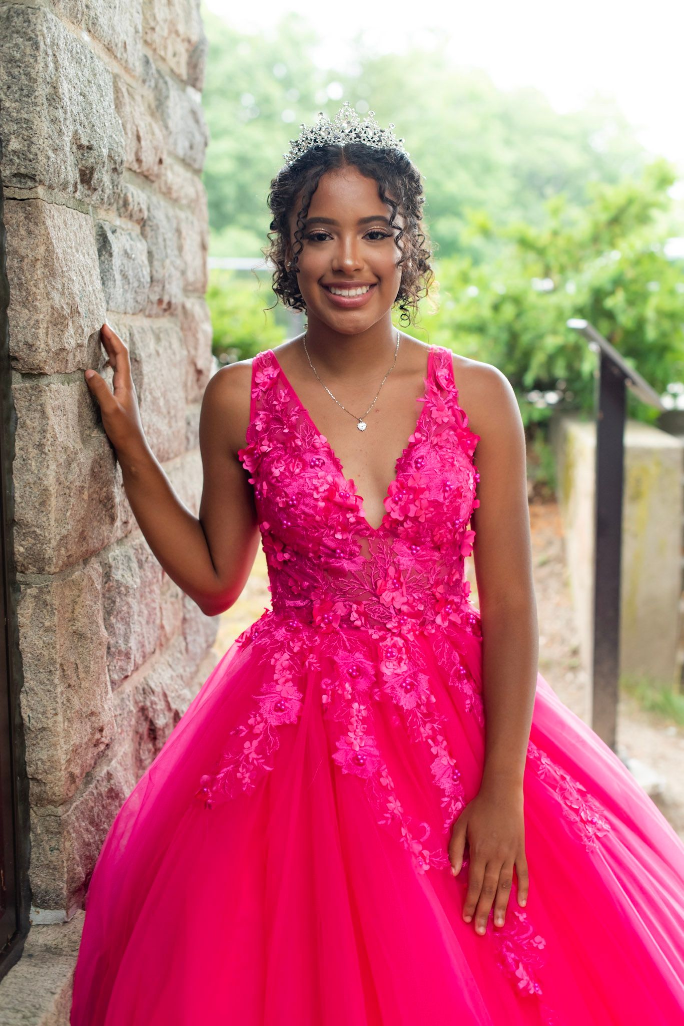 A person poses in a vibrant hot pink ball gown with lace details and a full tulle skirt against a stone wall backdrop.