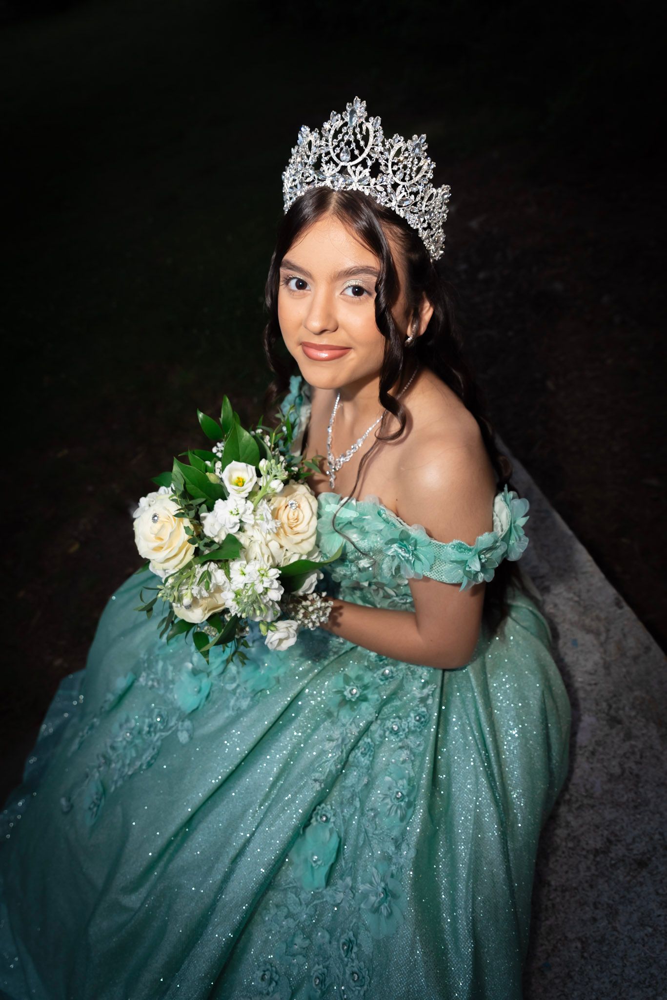 A quinceañera portrait session featuring a sparkling turquoise ball gown adorned with floral details and a crystal crown.