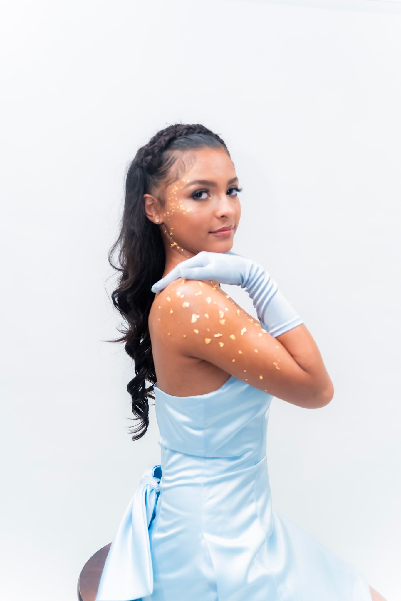 Woman in light blue dress posing against white background with sparkly body art details.