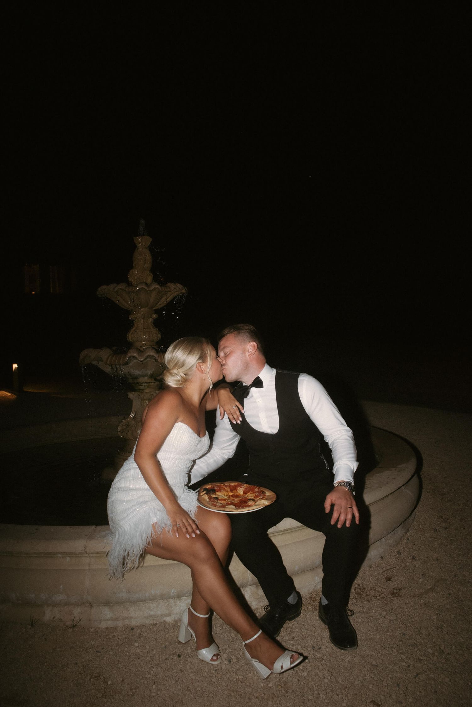 Newlyweds share a playful moment with pizza during nighttime wedding reception.