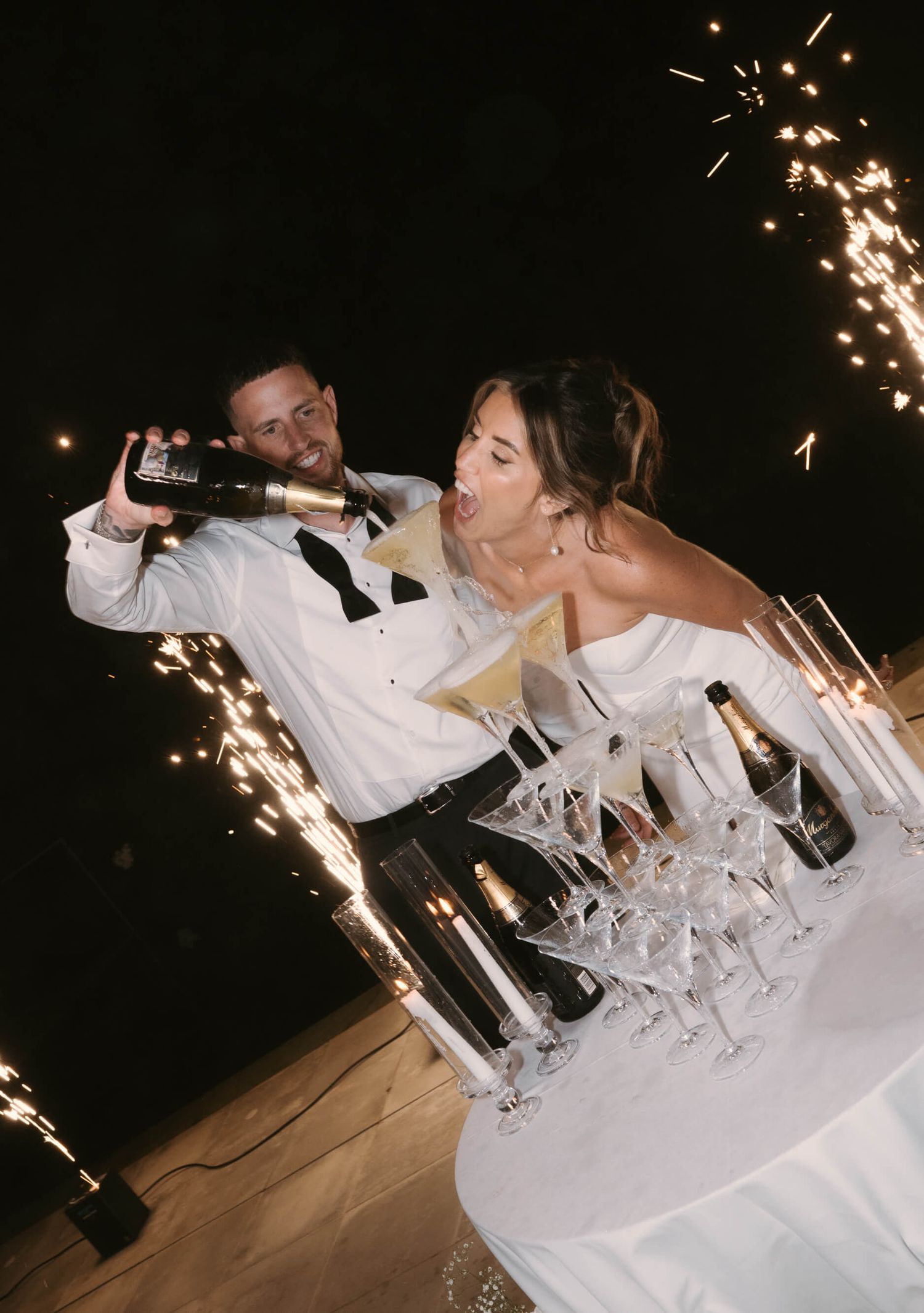 Wedding celebration with sparklers illuminating champagne tower at night.