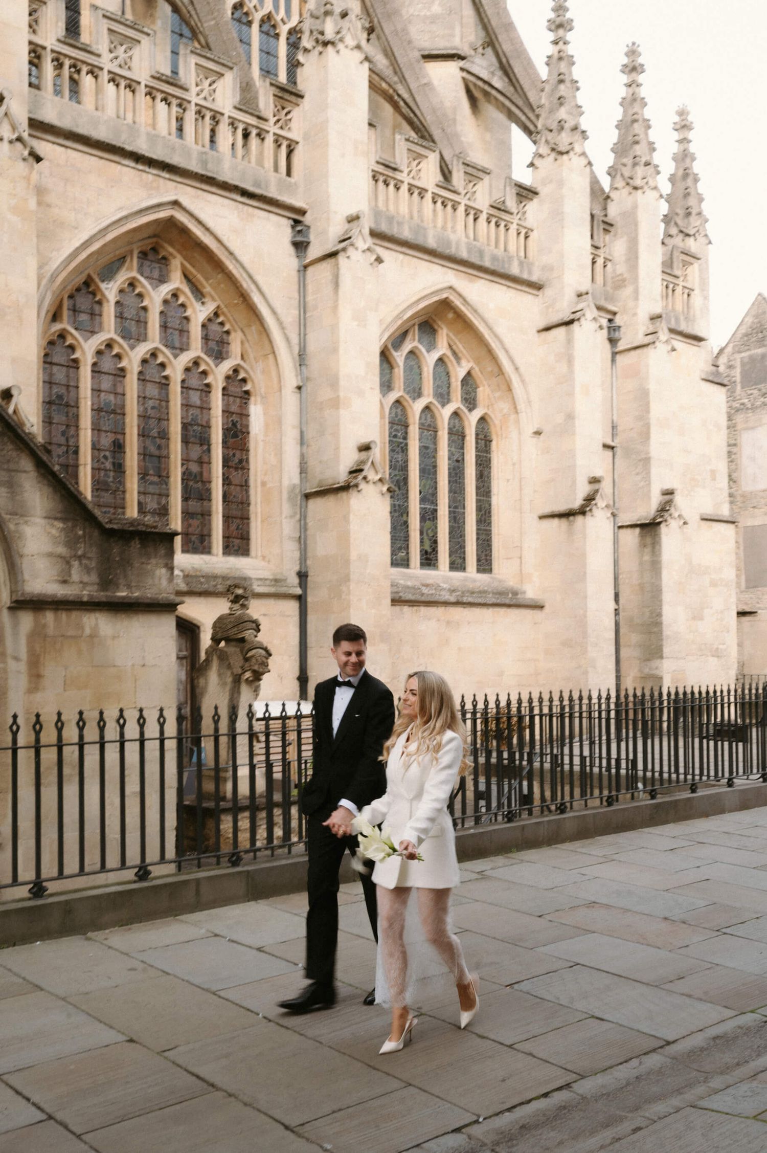 A couple walks along a historic stone cathedral exterior with Gothic architecture and ornate windows.