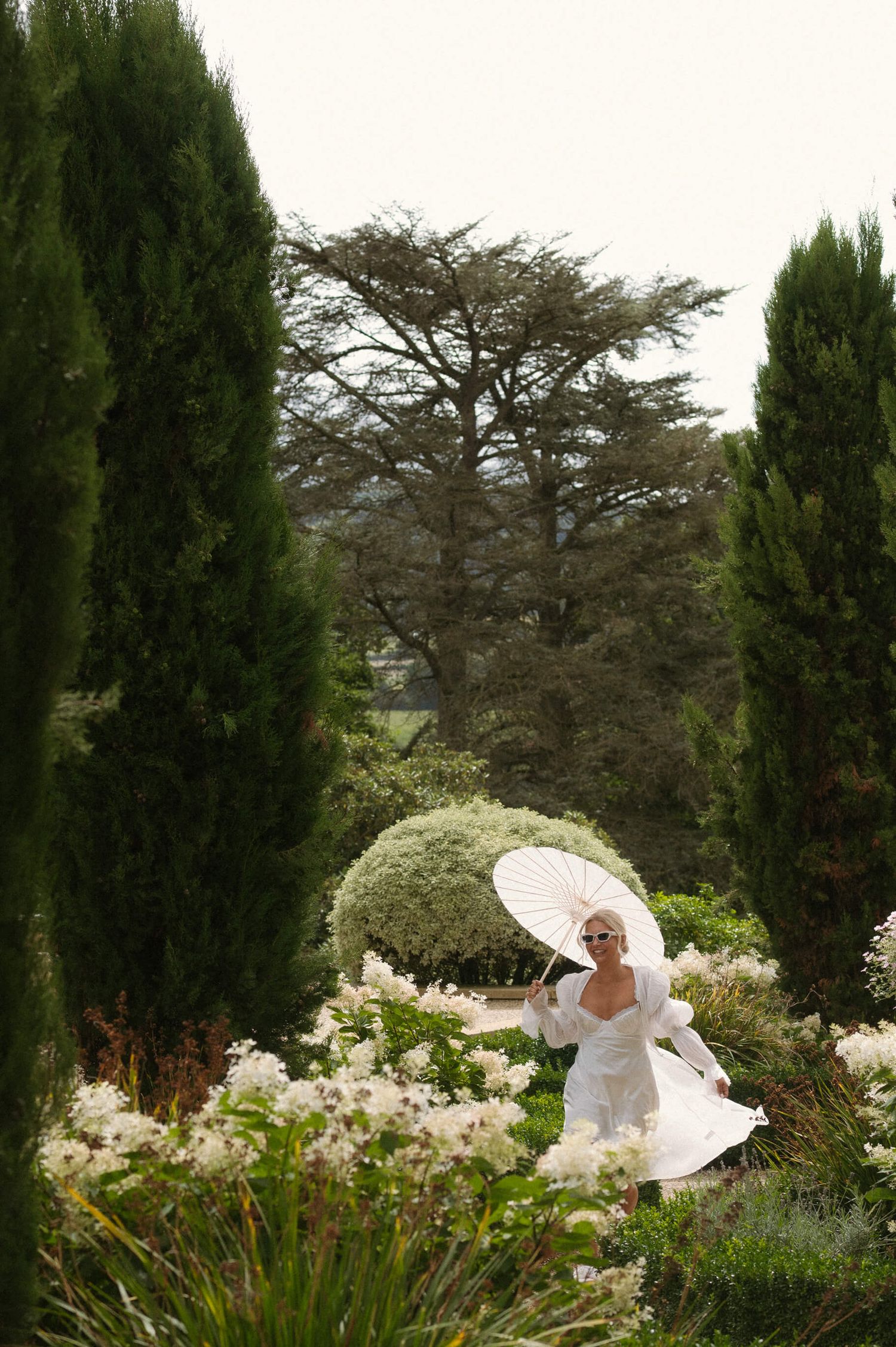 Figure in white dress with parasol poses among cypress trees in a manicured garden.