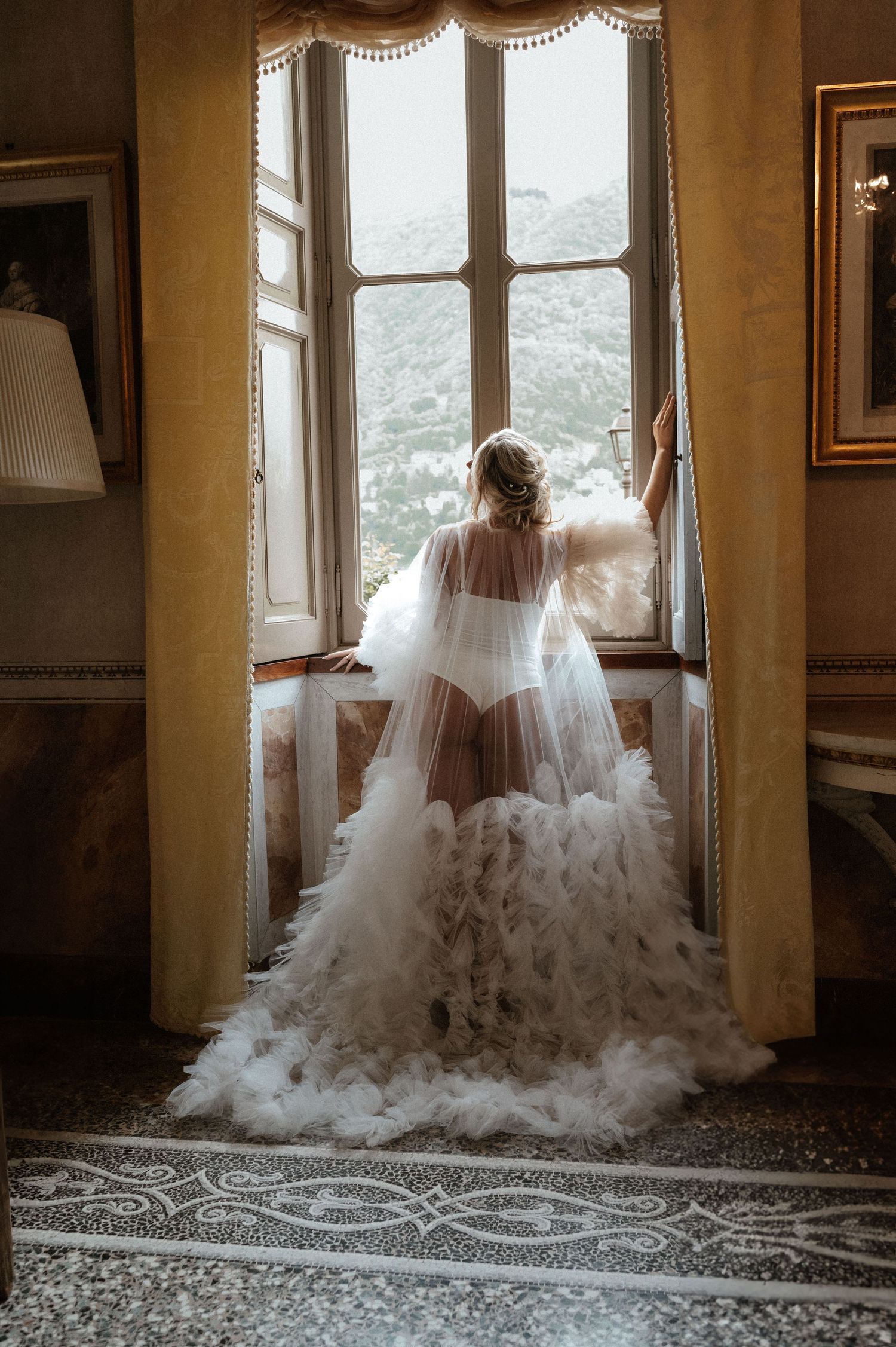 Ethereal bridal silhouette standing at an ornate window in a flowing tulle gown with mountain views.