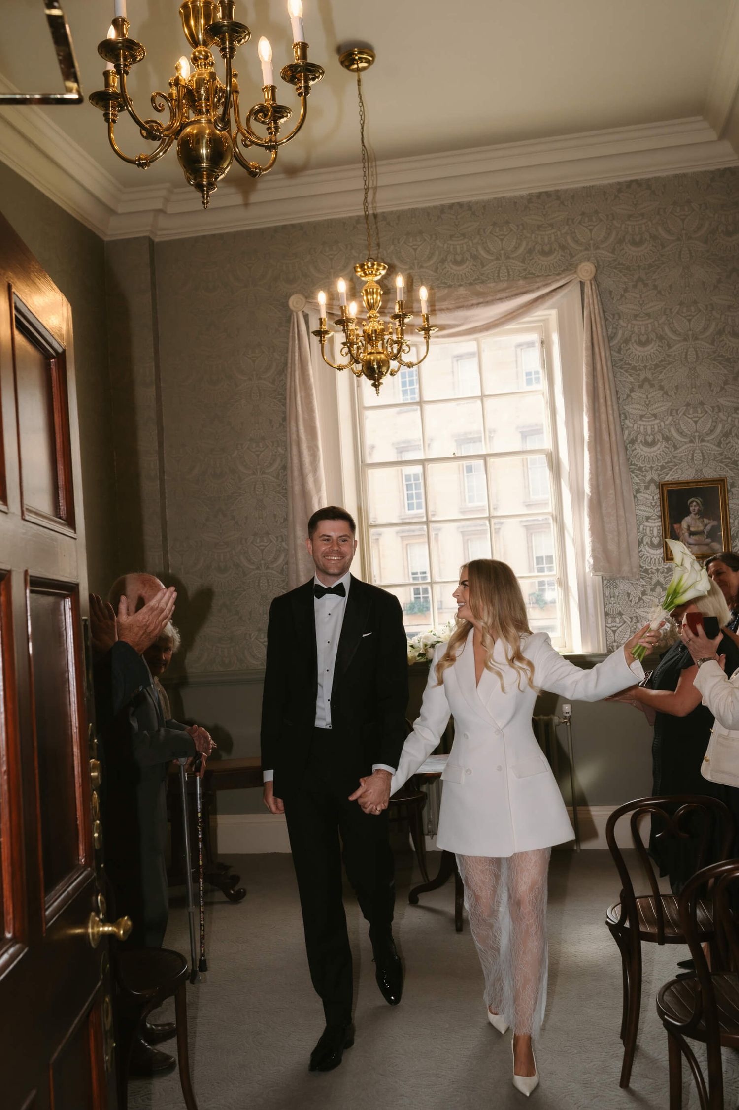 Newlyweds walking through an elegant room with ornate chandeliers and wallpaper.