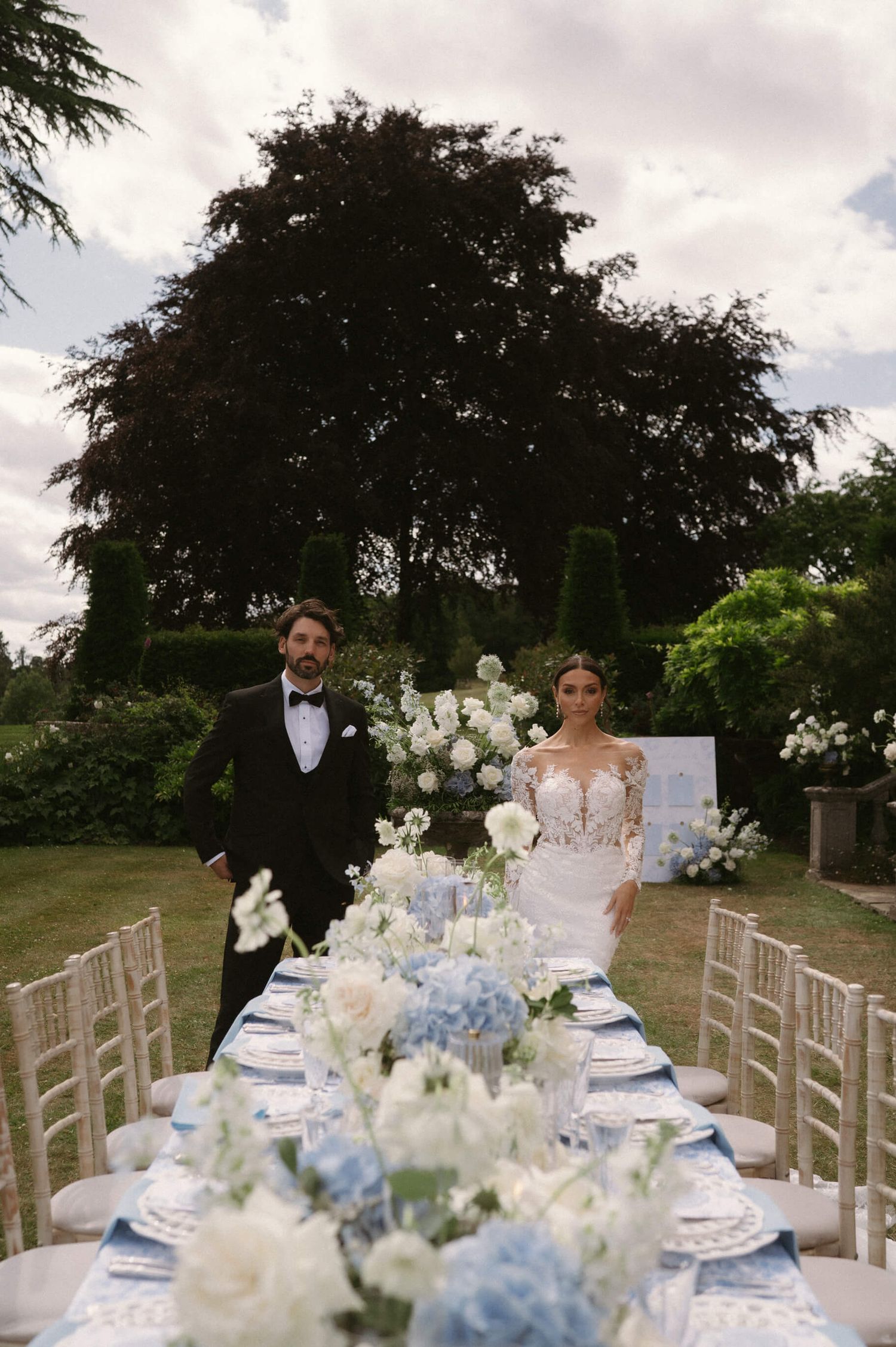 Outdoor wedding reception with blue and white table settings under trees in an elegant garden.