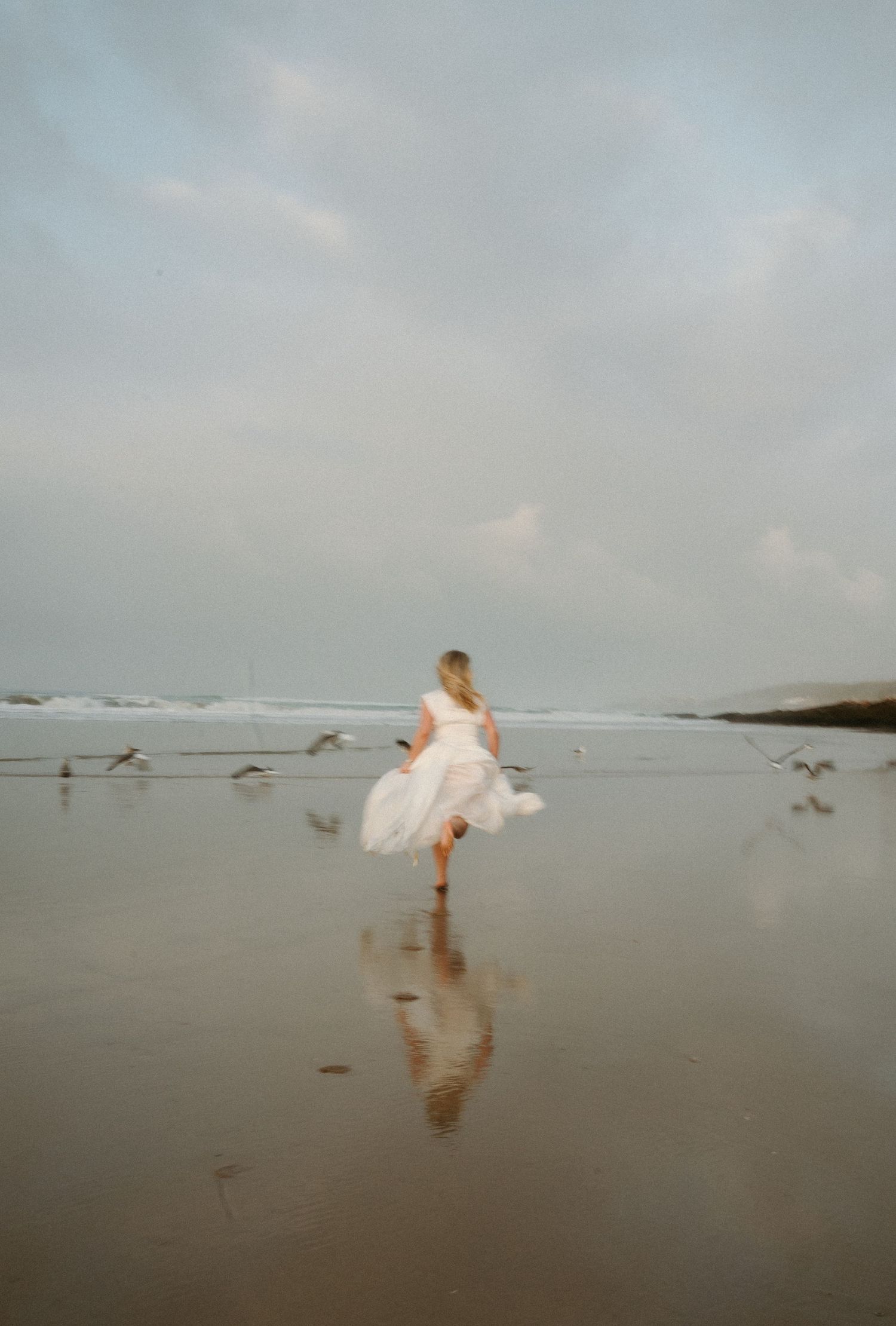 A figure in a flowing white dress runs along a misty beach shoreline at dusk creating a dreamy ethereal scene.