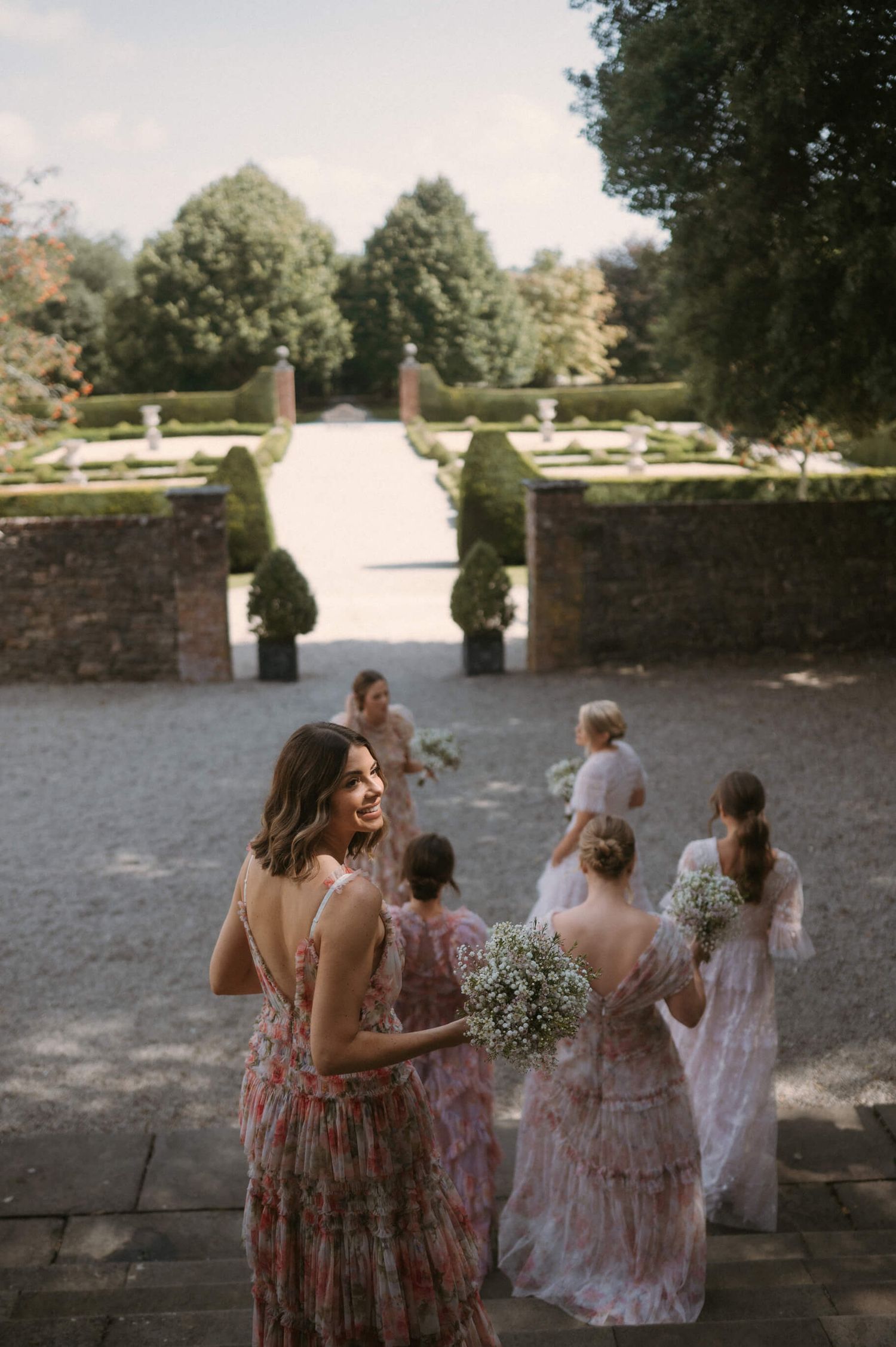 Wedding guests in floral dresses walk through a formal garden with manicured hedges.