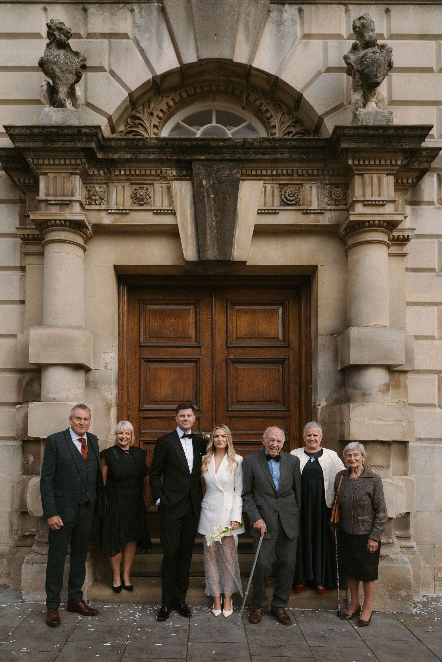 Group of formally dressed guests stand outside ornate wooden church doors for a special occasion.
