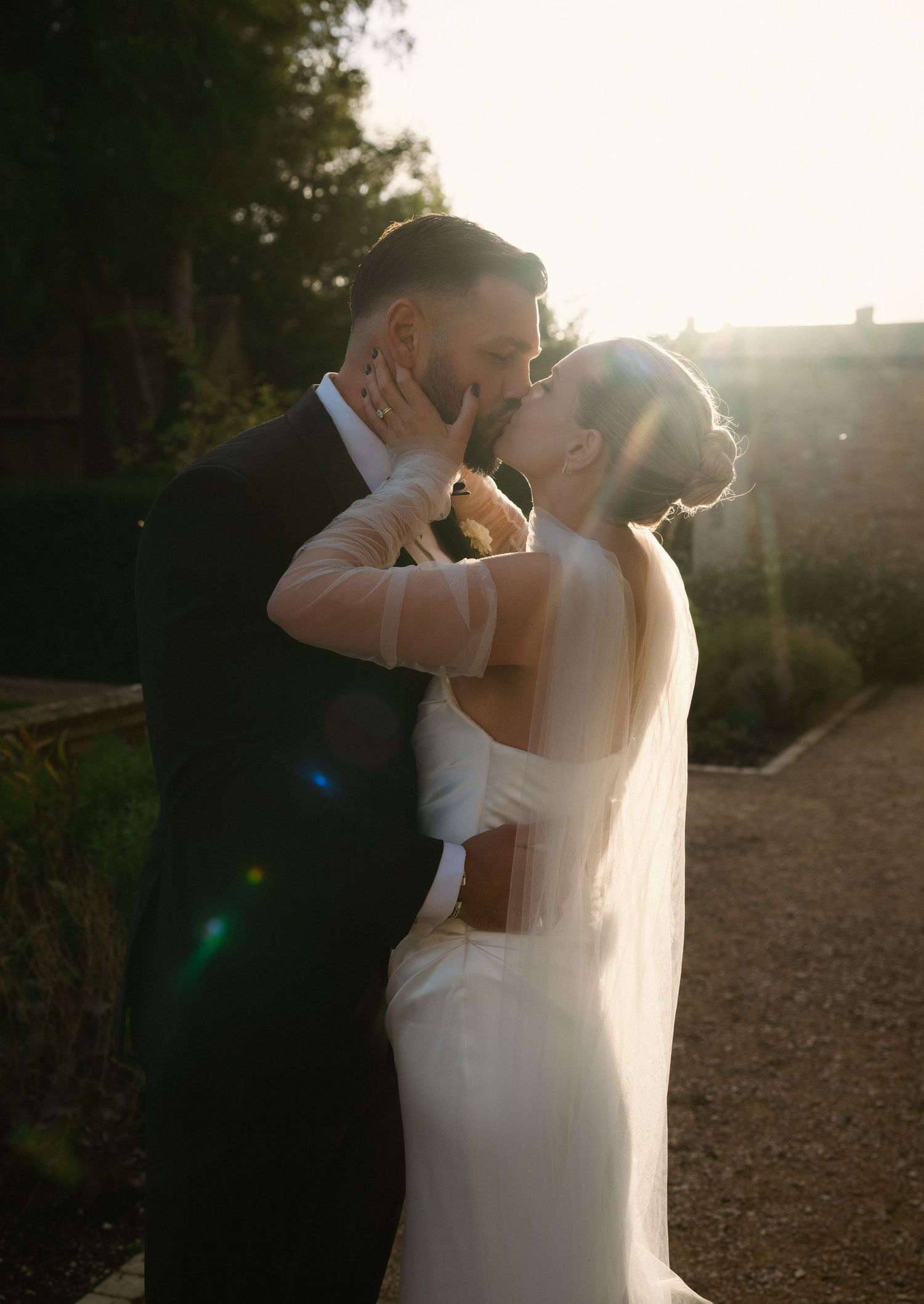 Bride in a white satin gown and veil kisses her groom in a dark suit in a sunlit garden during golden hour.