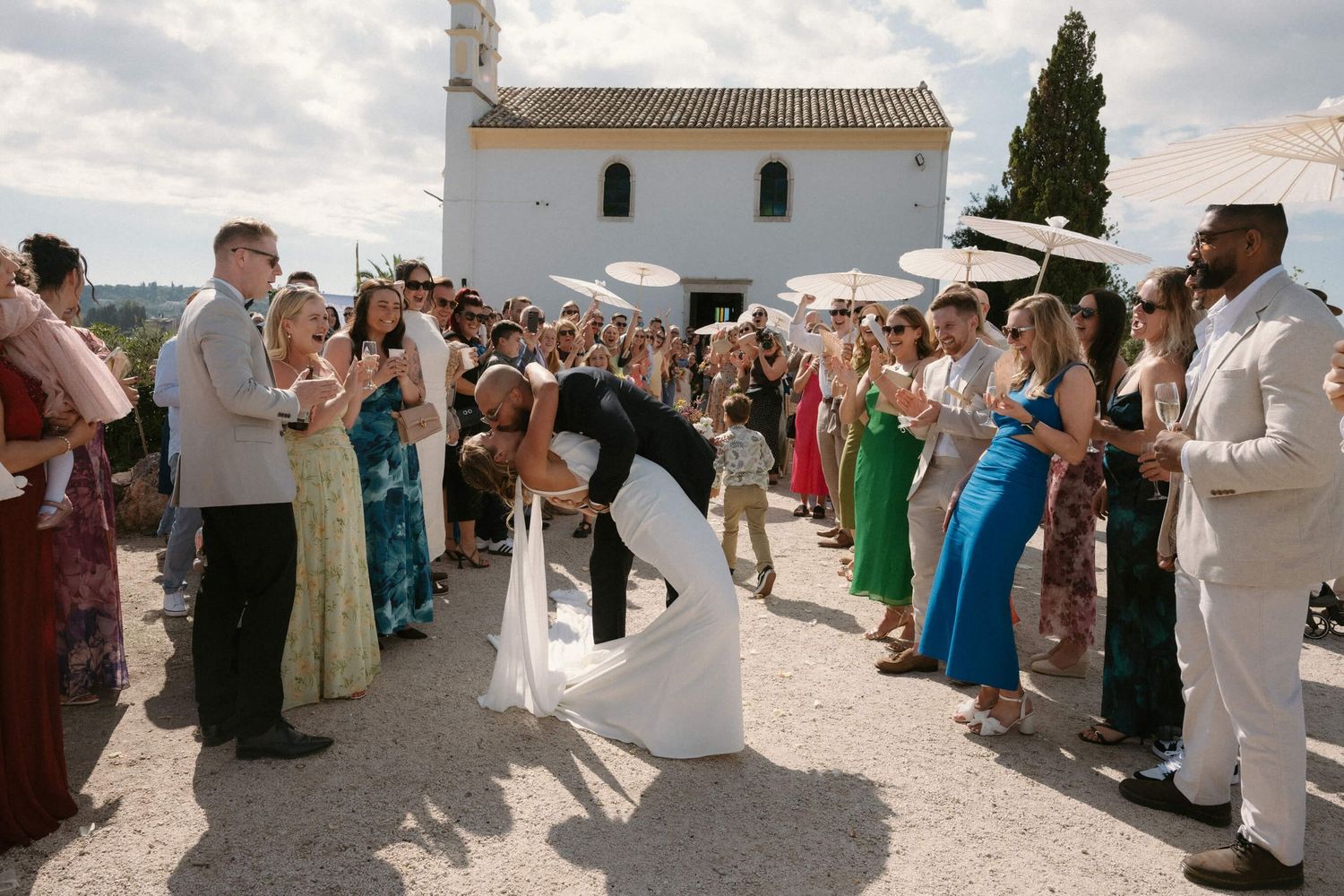 Wedding ceremony exit with guests forming an aisle outside a white Mediterranean chapel.