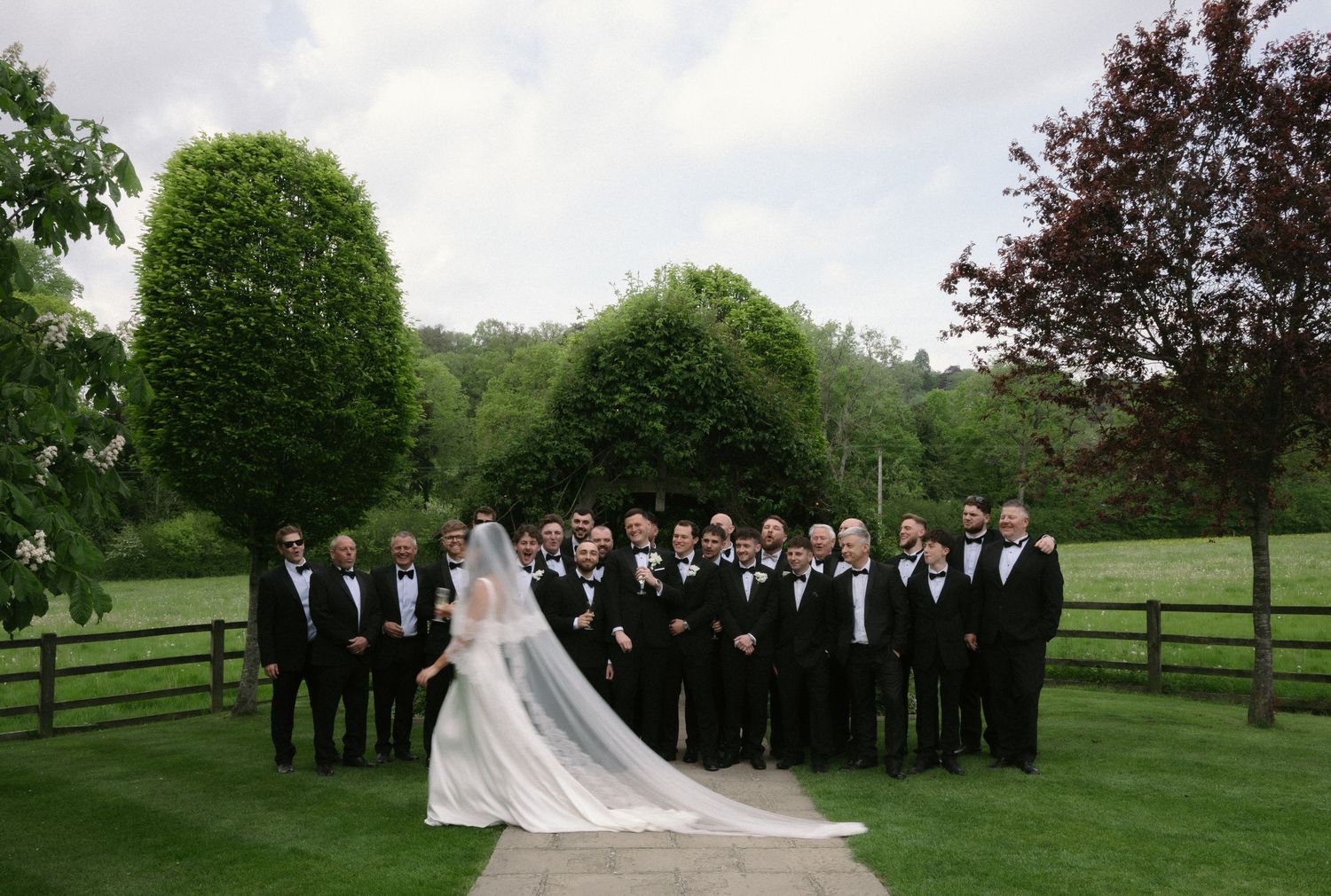 Bride in a long white gown and veil stands before a large group of groomsmen in black tuxedos on a green lawn at an outdoor wedding.