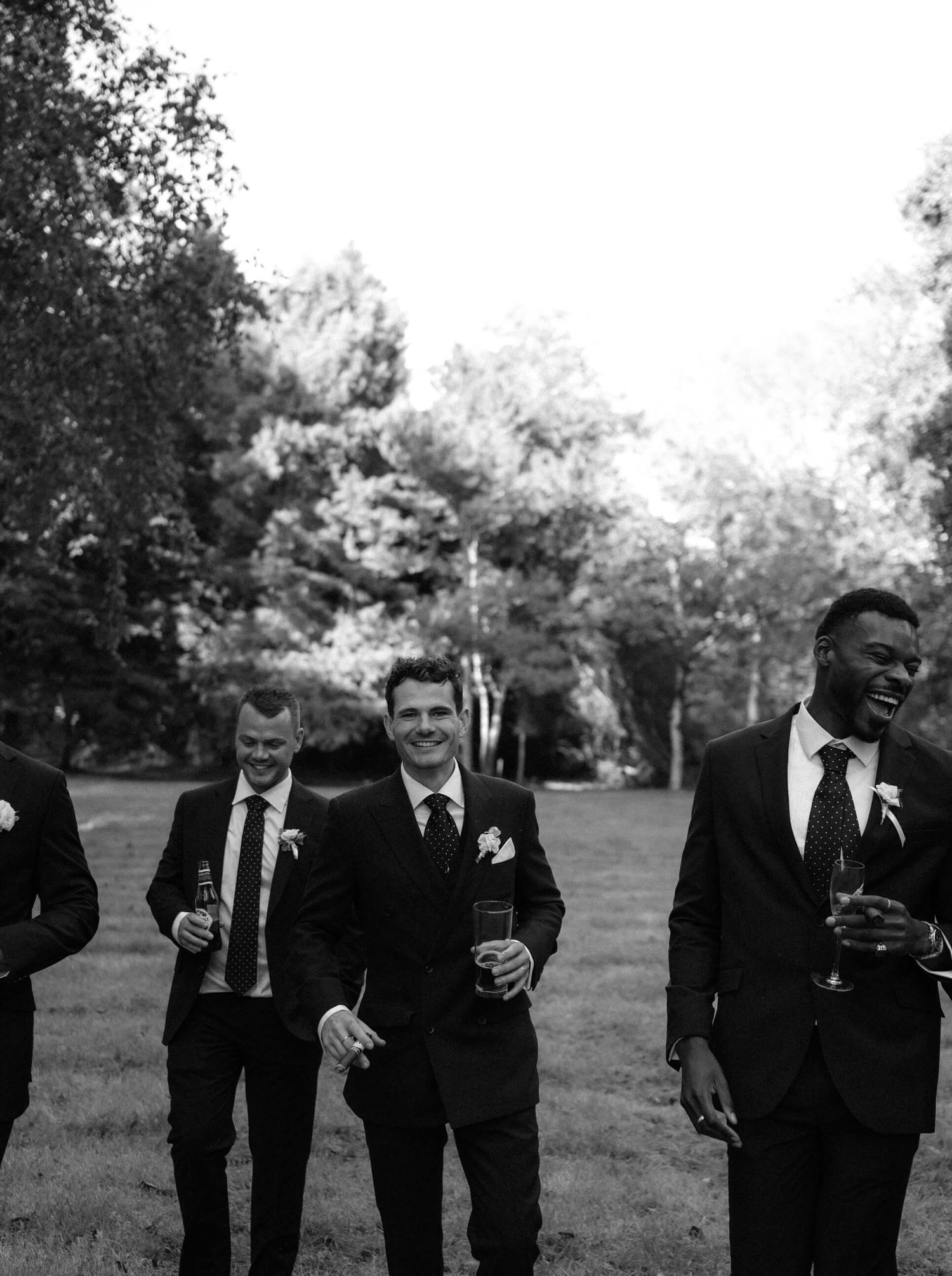 Group of groomsmen in black suits walking together across a grassy outdoor setting.