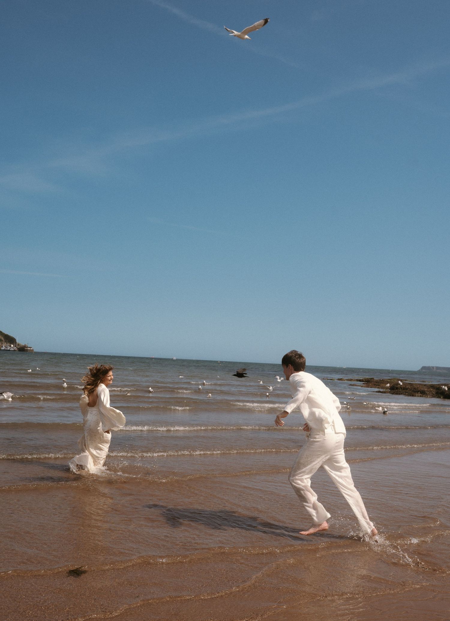 A couple in white outfits playfully run along a beach shore as a seabird soars overhead.