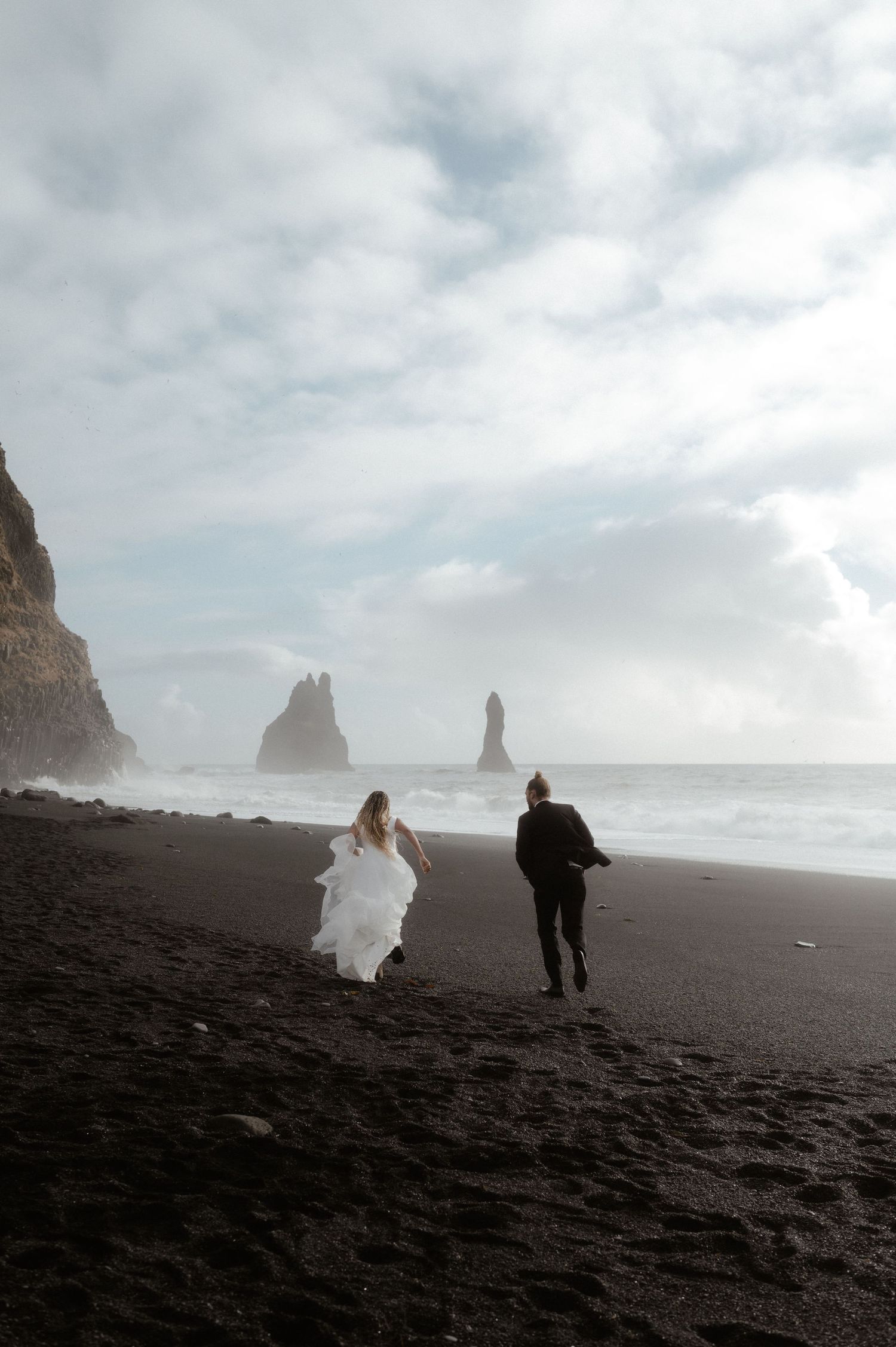 Two people run along a dramatic black sand beach with towering sea stacks and misty cliffs in Iceland.