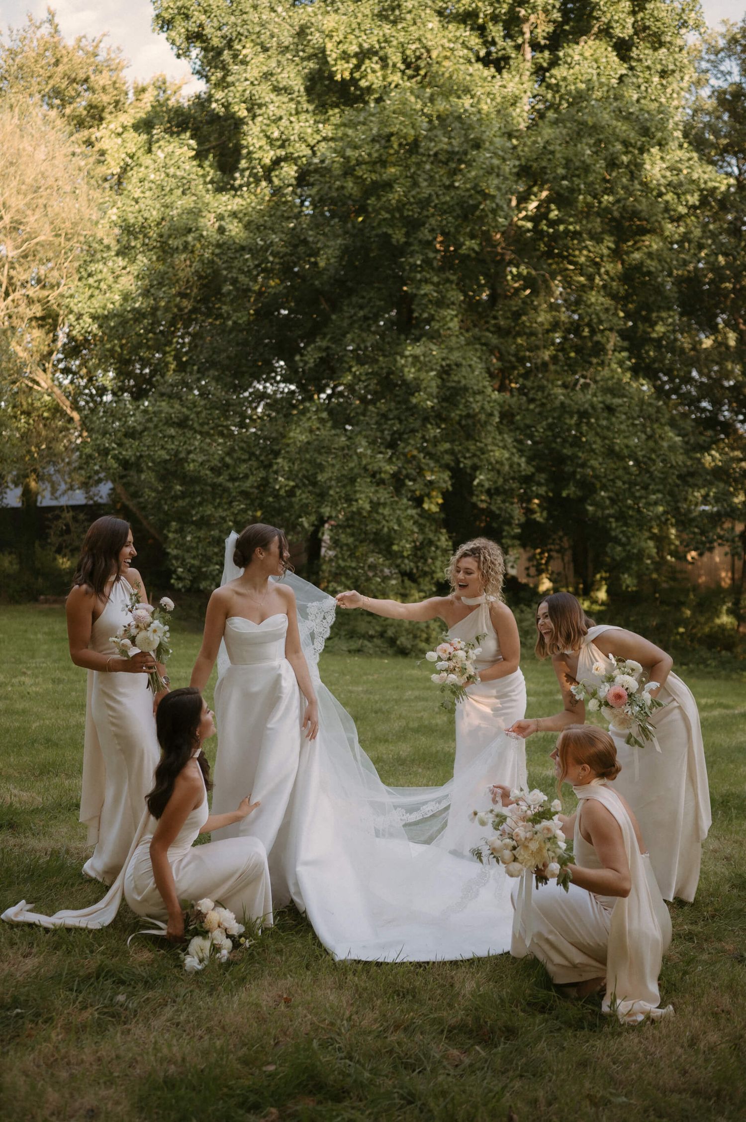 Bridesmaids in light dresses help arrange the bride's wedding gown train on a sunny lawn.