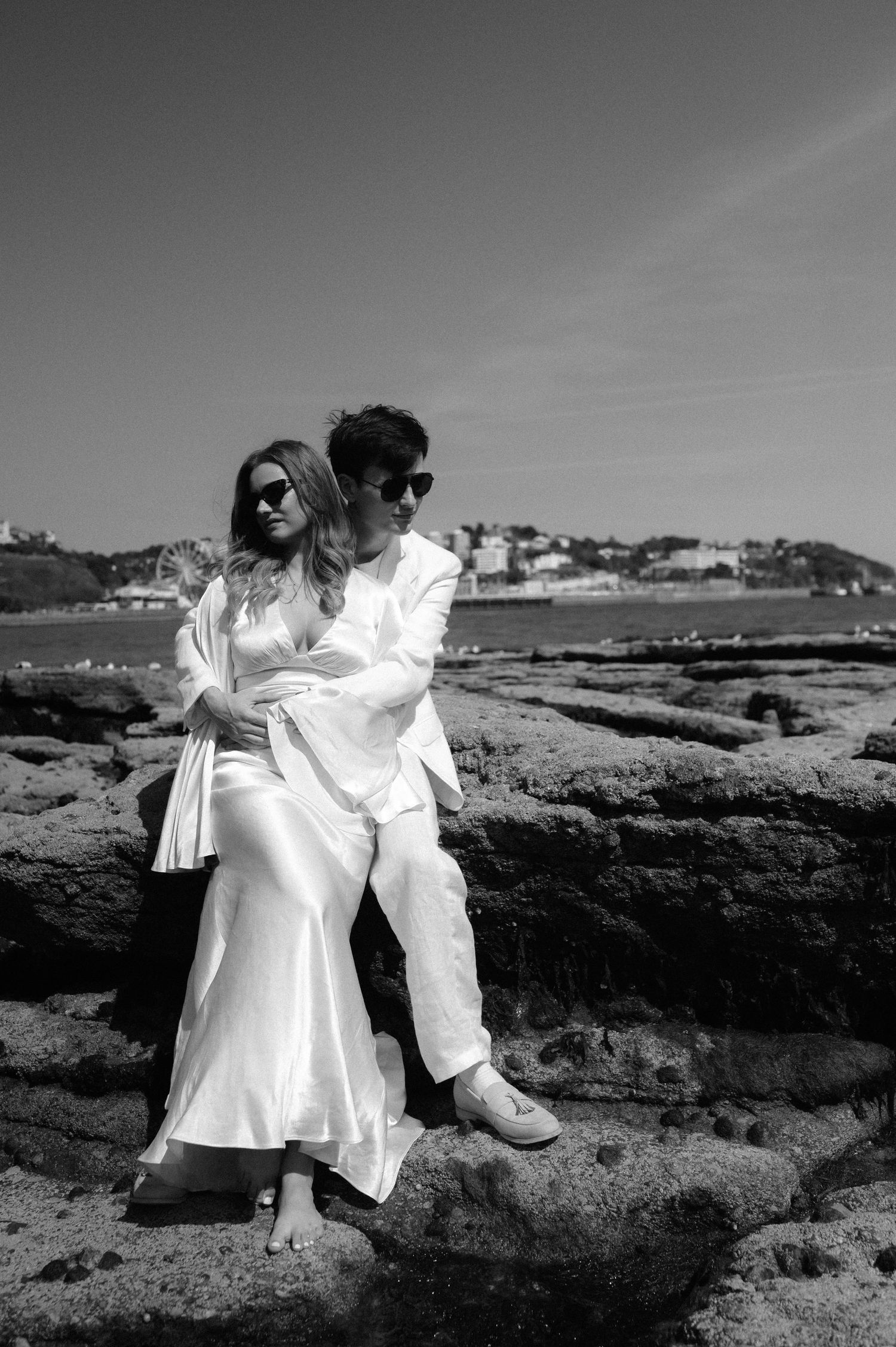 A romantic black and white photo of a couple in white clothing embracing on coastal rocks.