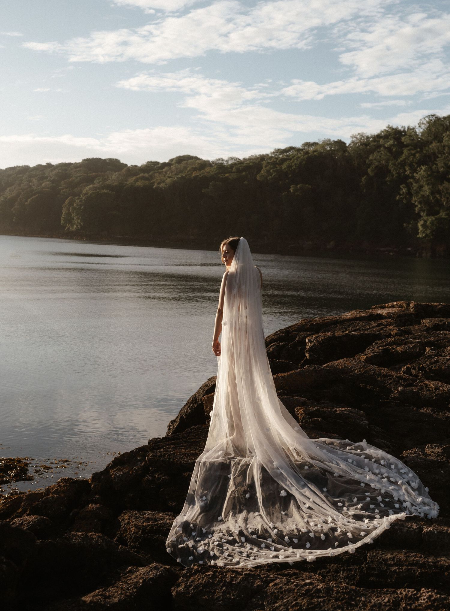 A dramatic bridal portrait shows a flowing lace veil trailing over rocky cliffs beside a serene lake at sunset.