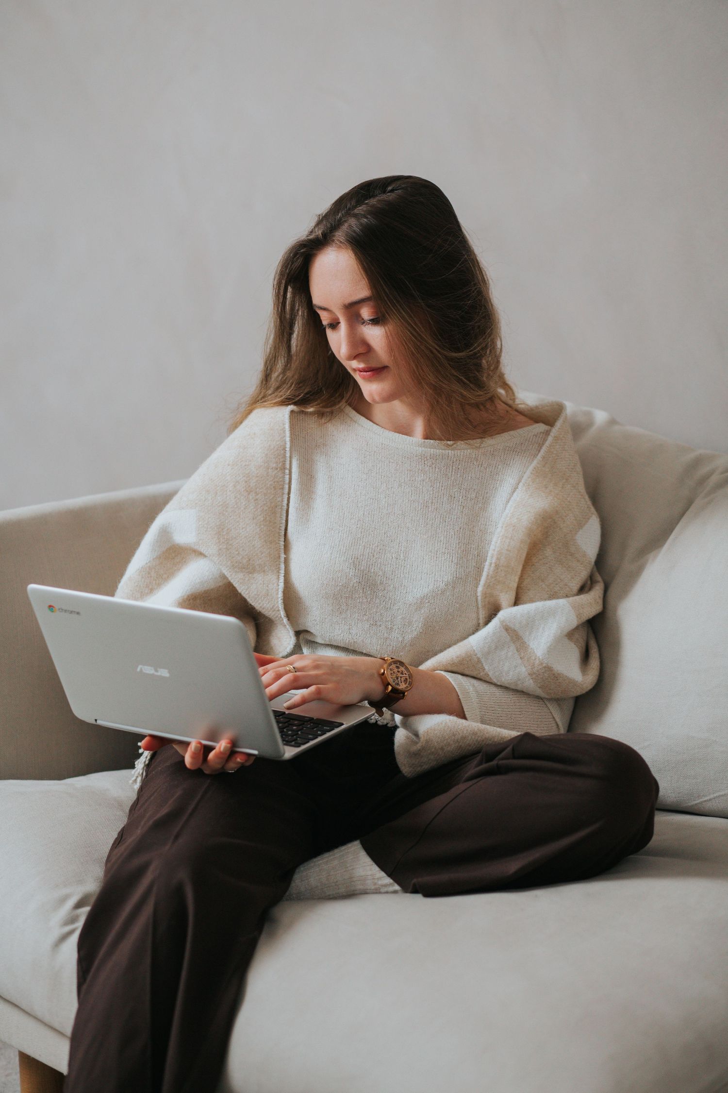 Person in cream sweater relaxing on white couch while using laptop computer.