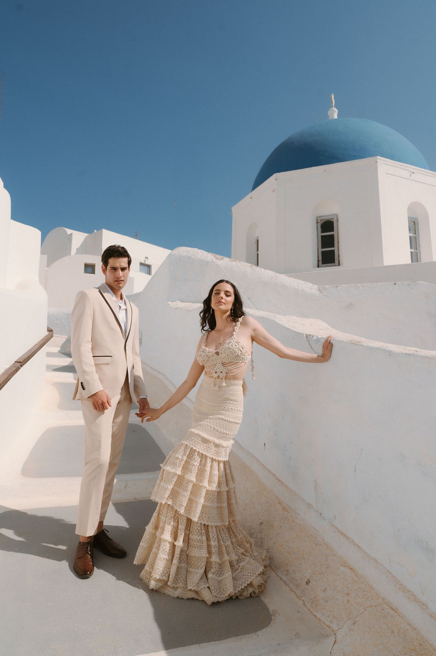 Couple in elegant cream attire pose on whitewashed steps beside a blue-domed church in Santorini, Greece, under a clear sky.