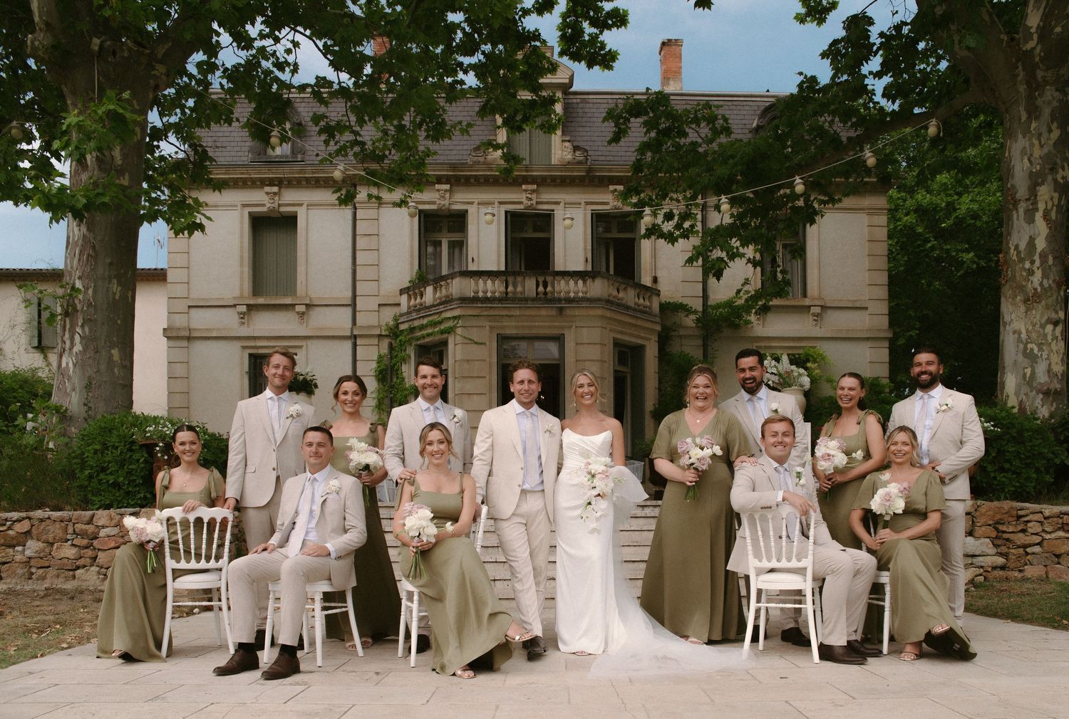 A panoramic sequence of wedding party members in white attire sitting on chairs along a stone wall outdoors.