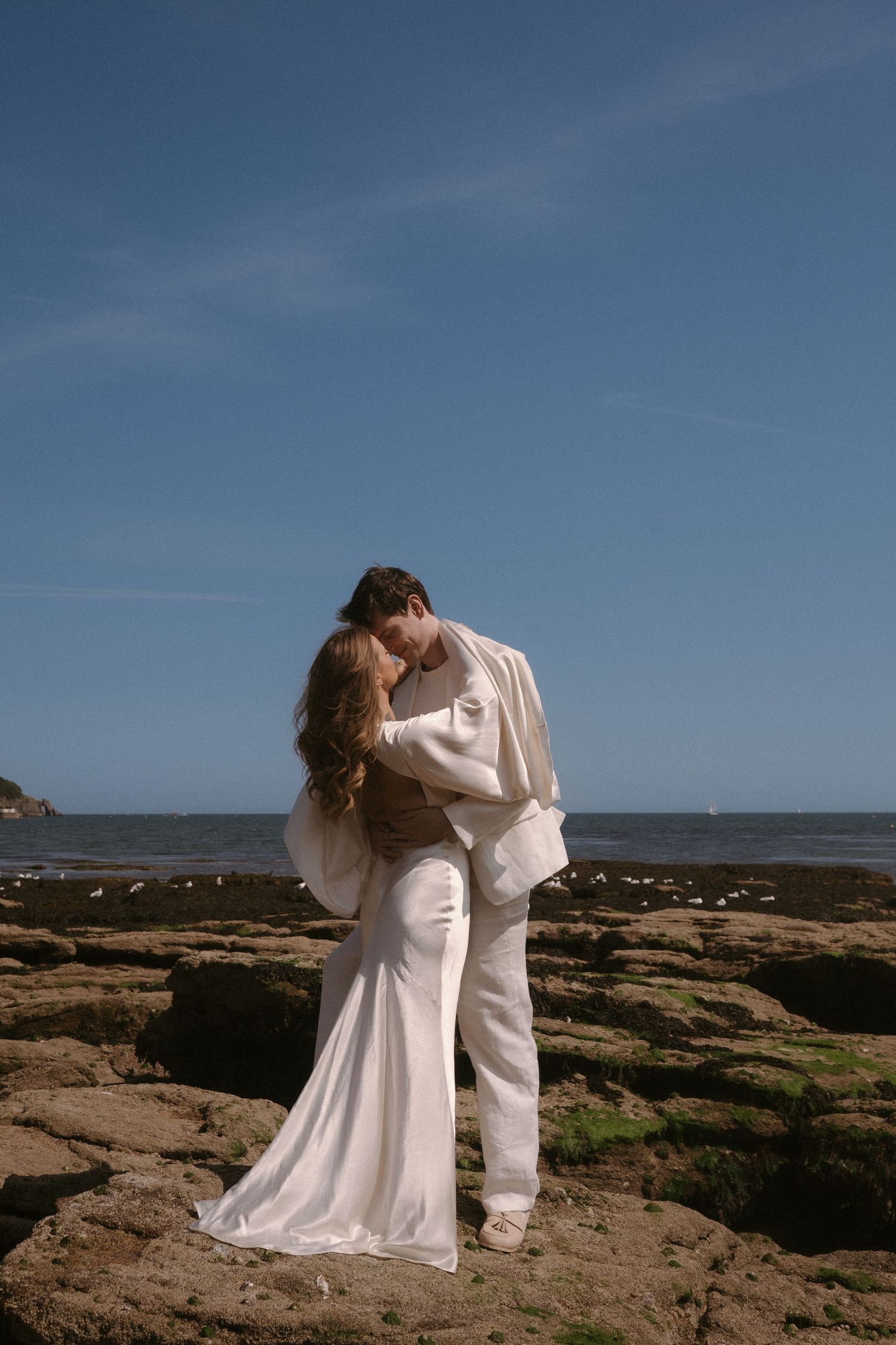Couple in flowing white outfits embracing on rocky coastal cliffs under blue sky.