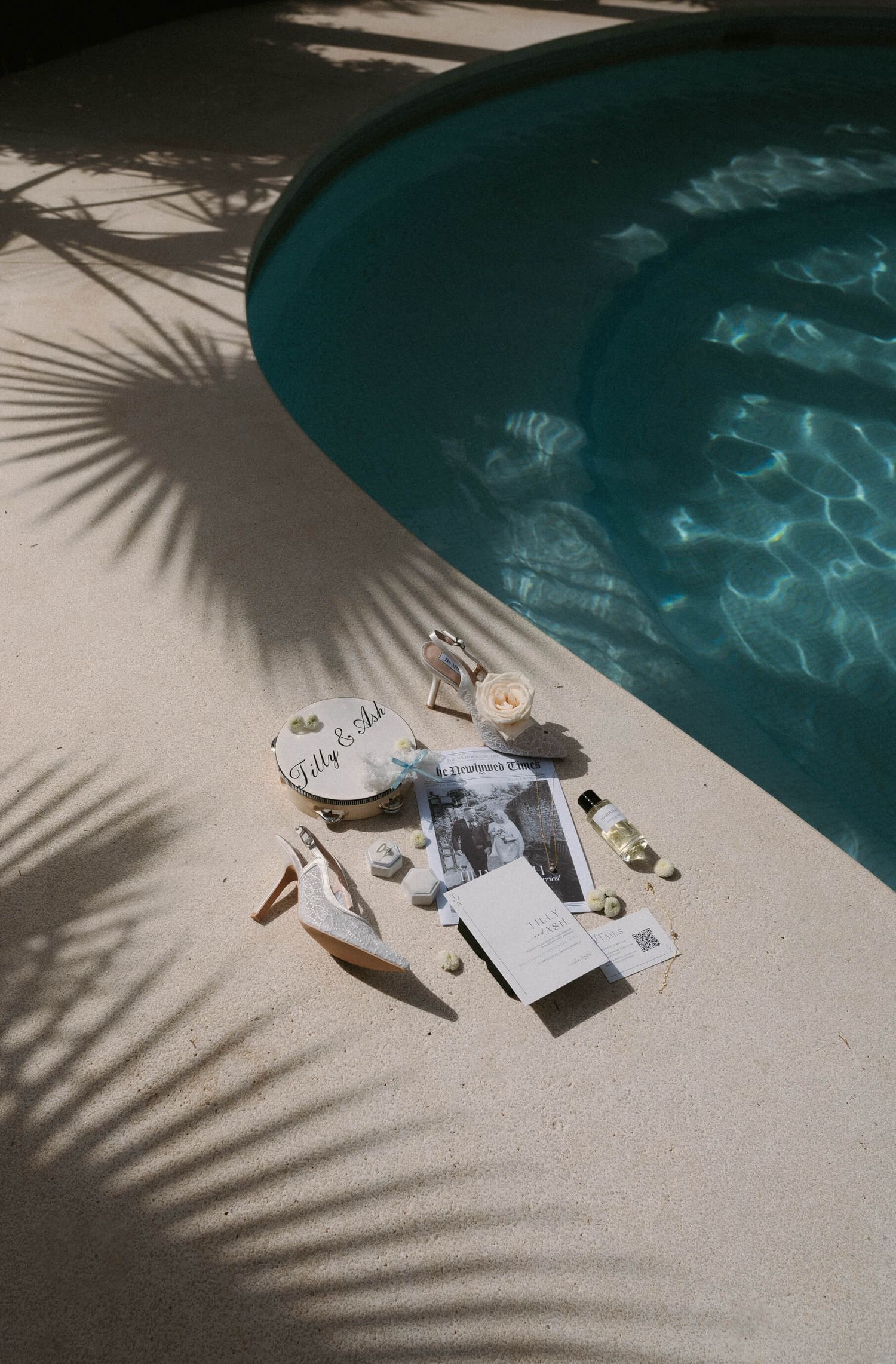 Wedding stationery and photographs artistically arranged by a pool with palm tree shadows.