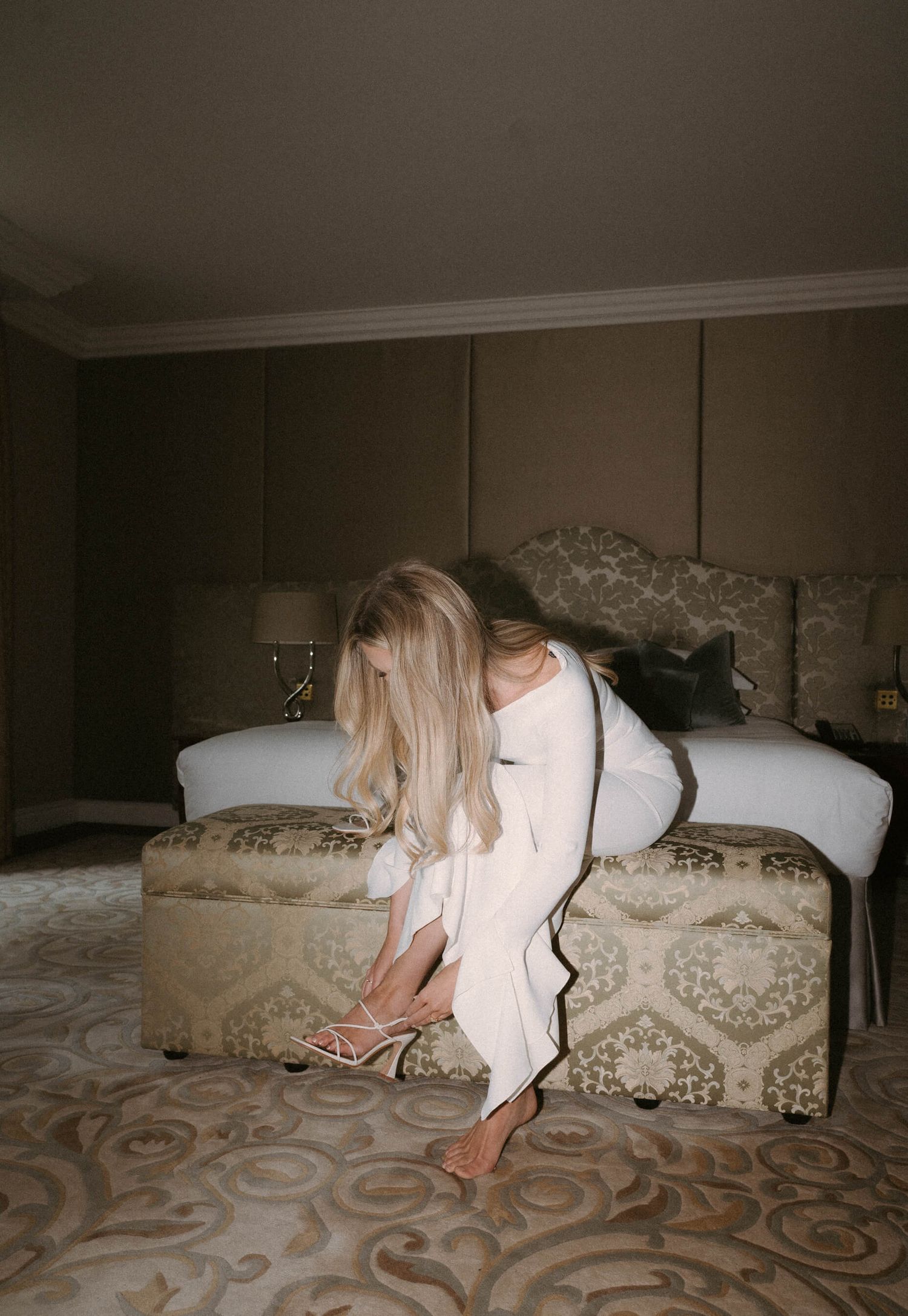 Person in white dress sitting on an ornate bed in a dimly lit hotel room.