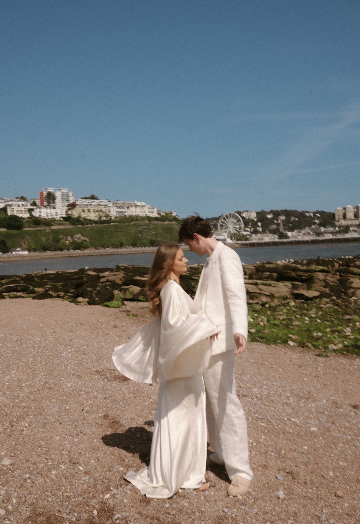 Couple in wedding attire sharing romantic moments on a rocky beach with ocean and buildings in background.