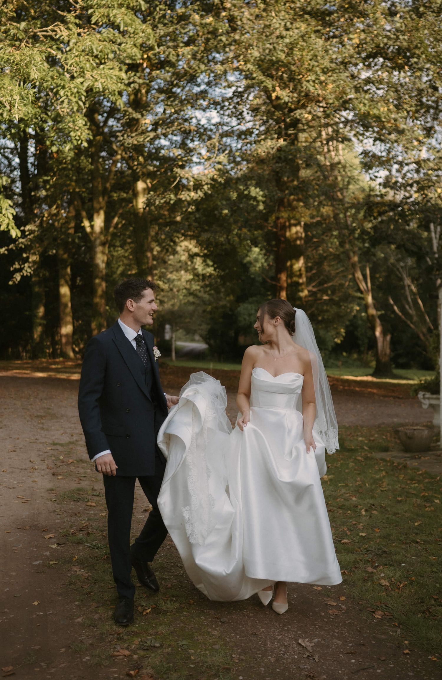 Wedding couple playfully walking through an autumn garden path with flowing white dress.