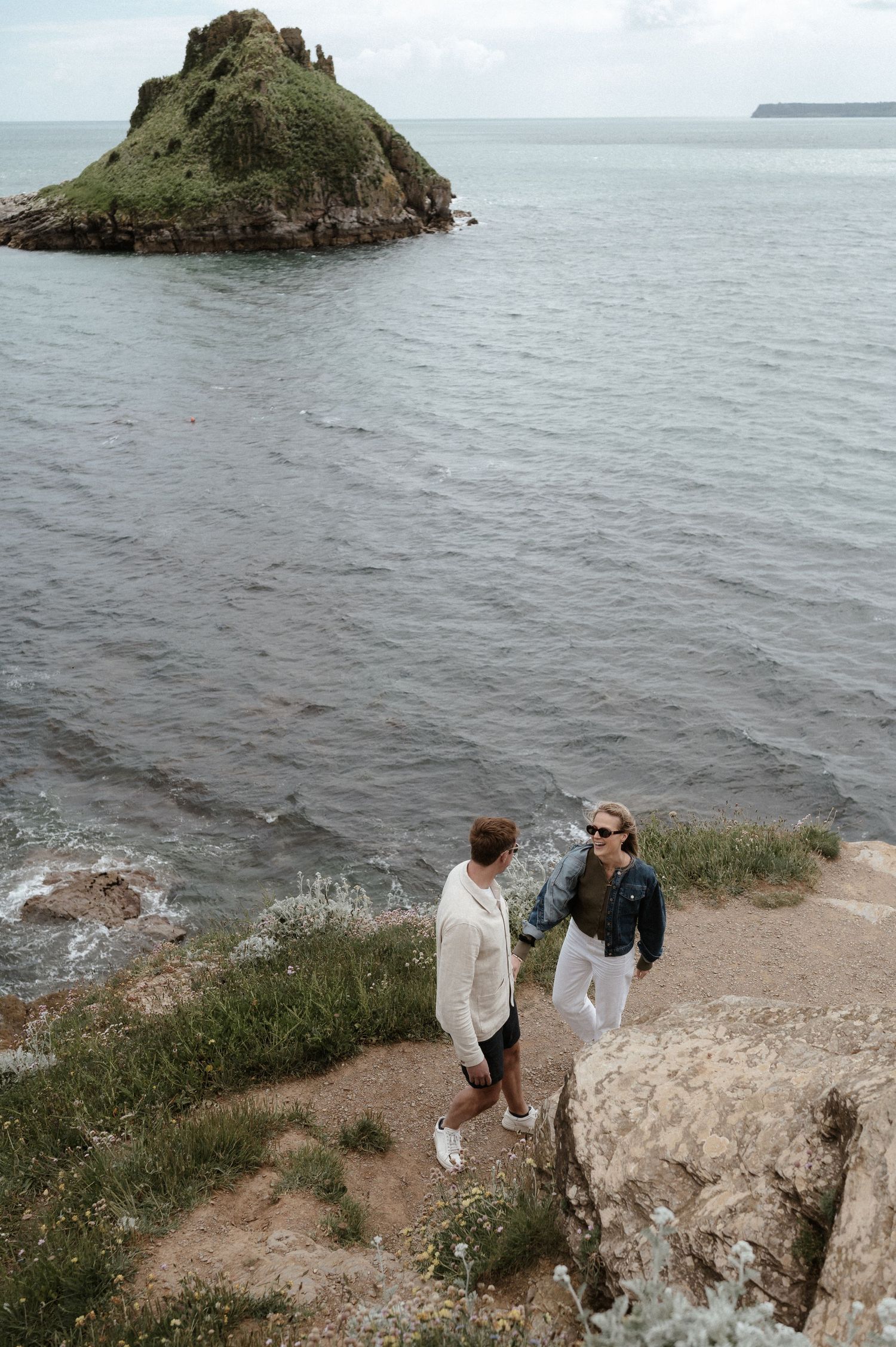 Couple stands on coastal cliff path overlooking dramatic rocky island formation.