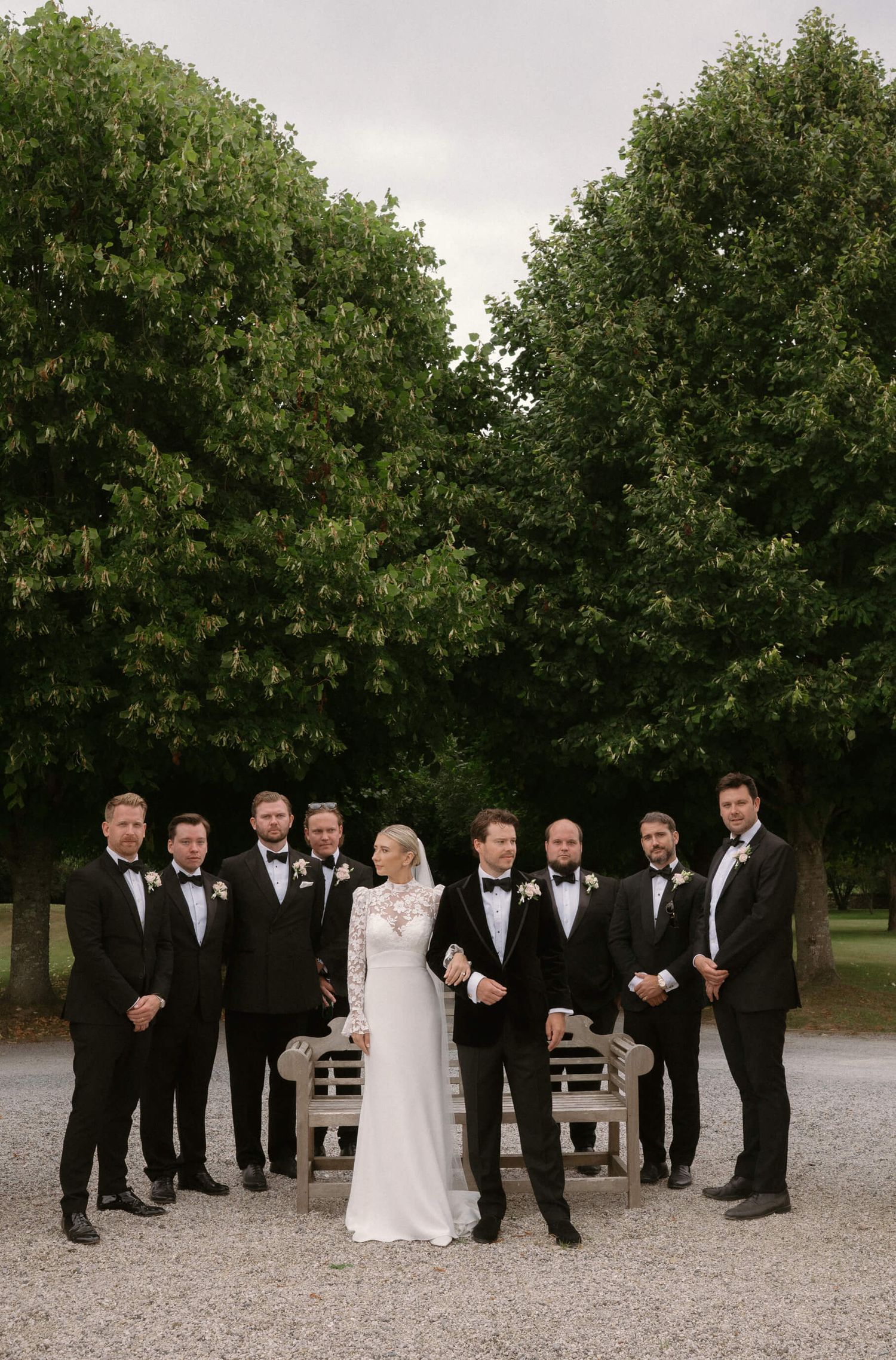 Wedding party in black tuxedos and white dress pose together on a gravel path surrounded by trees.