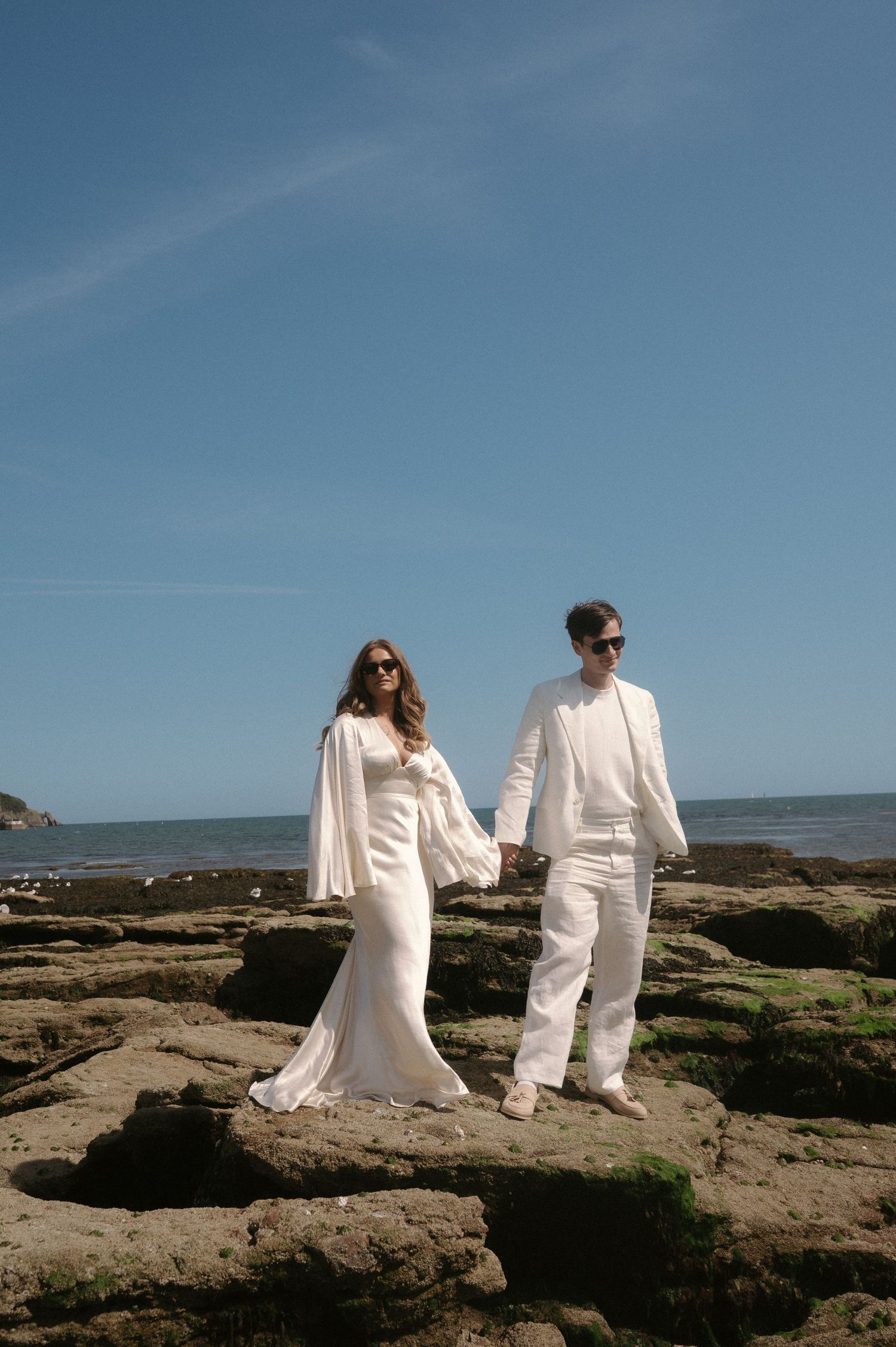 A couple in matching white outfits holds hands while standing on rocky coastal terrain against a blue sky.