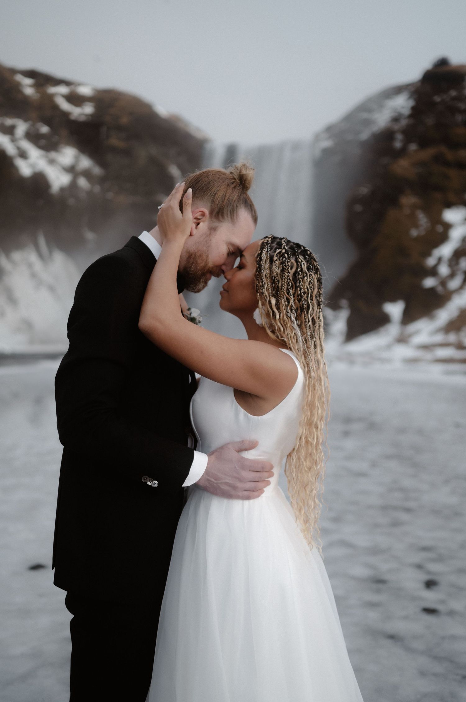 A couple shares a romantic kiss in front of a misty waterfall backdrop while dressed in formal wedding attire.