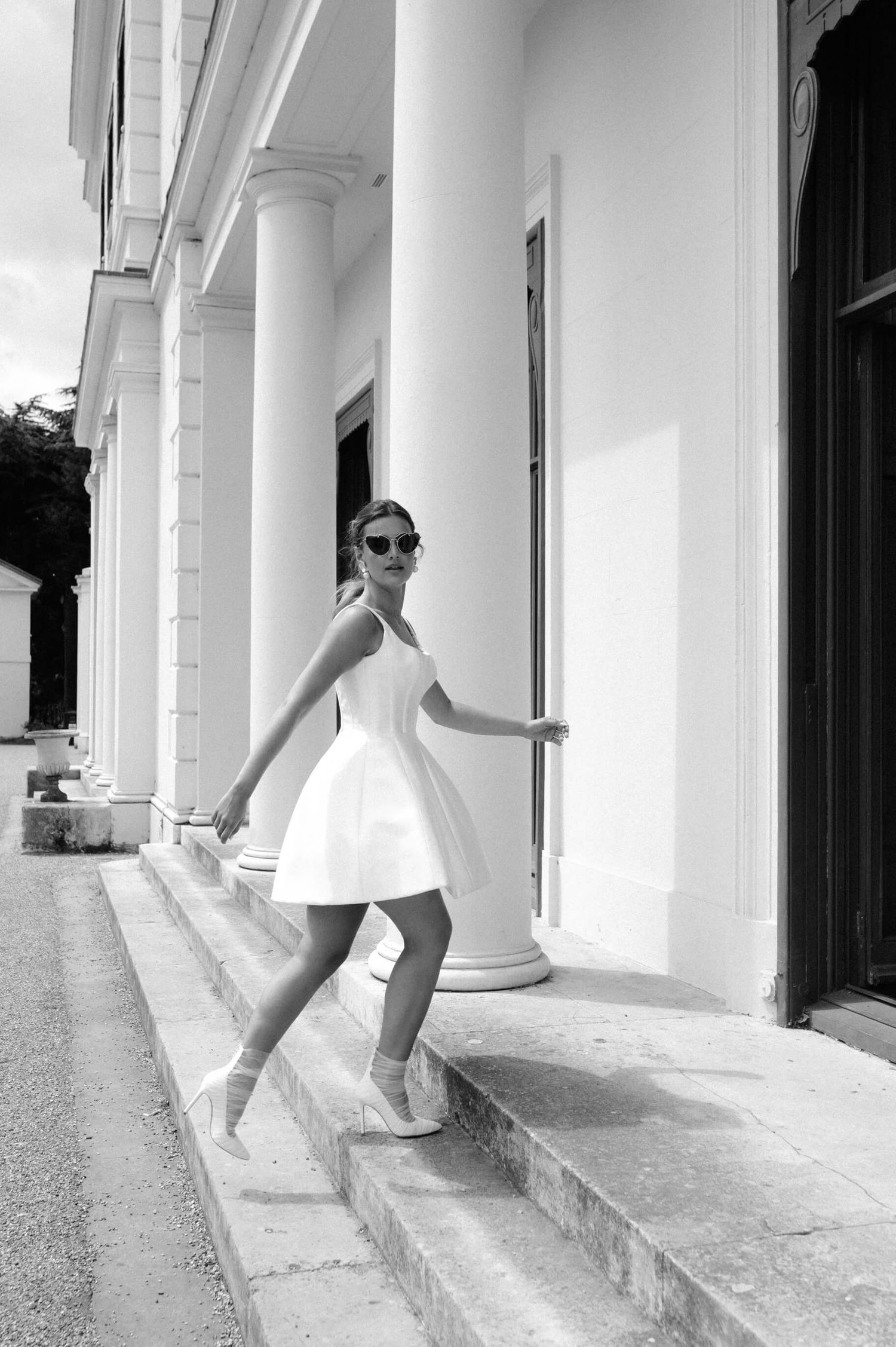 Bride in sunglasses poses playfully along a white columned building walkway.