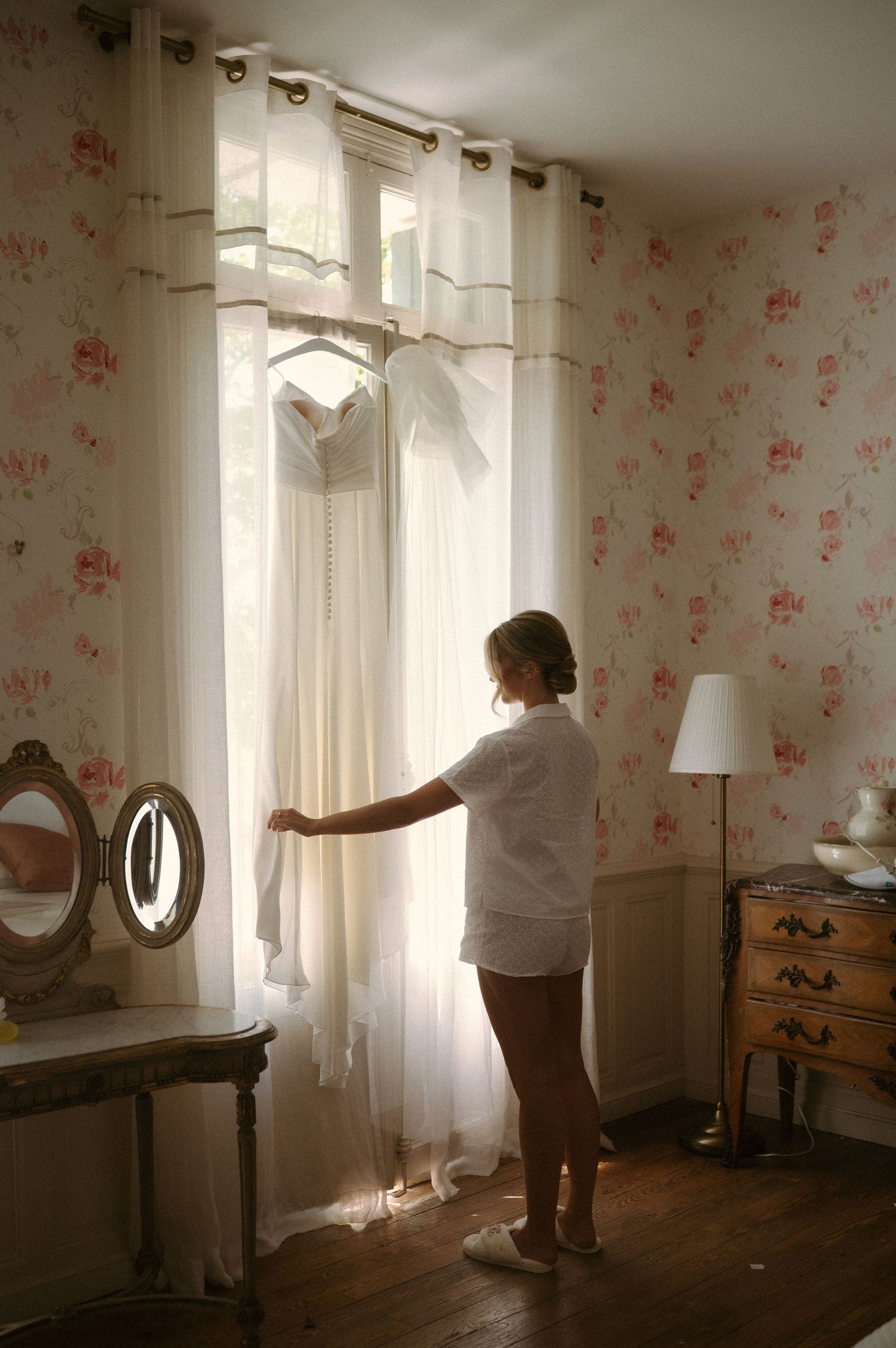 A person adjusts sheer white curtains in a sunlit bedroom with floral wallpaper and vintage wooden furniture.