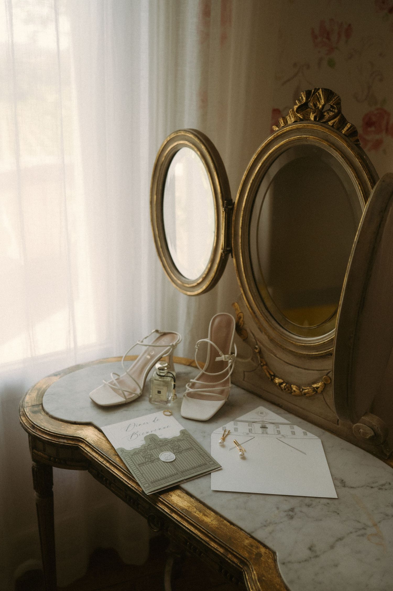 Vintage white shoes and a golden ornate mirror on a marble vanity table beside a sunlit window.