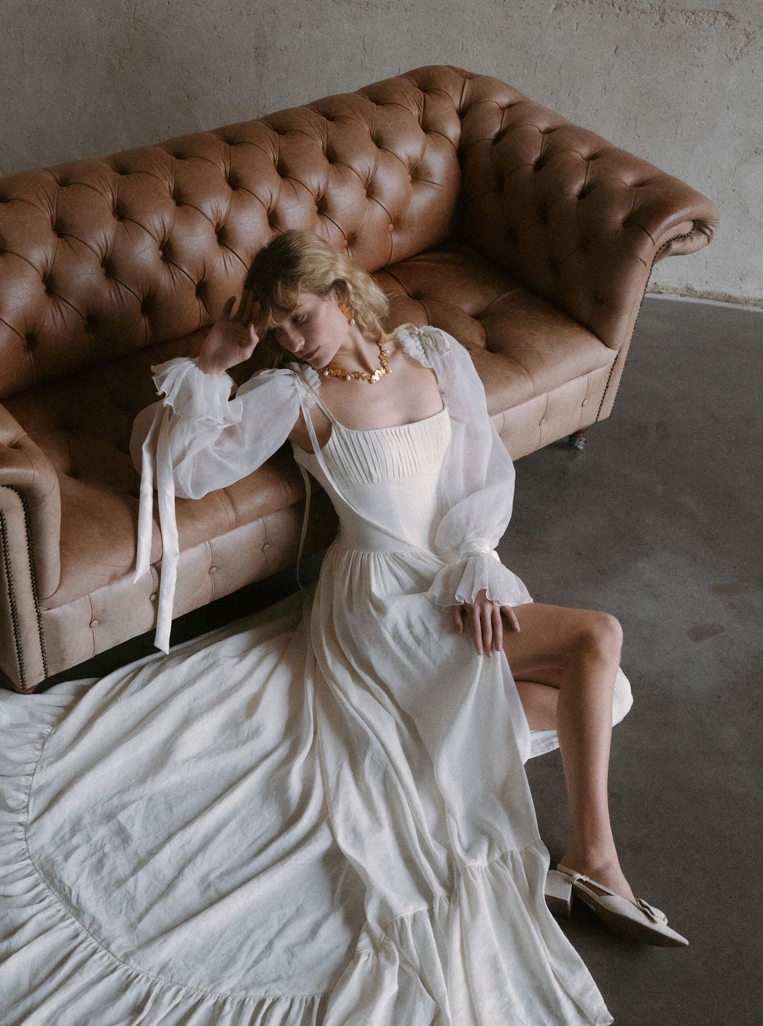 Elegant white wedding dress spread across a brown tufted leather couch in an industrial space.