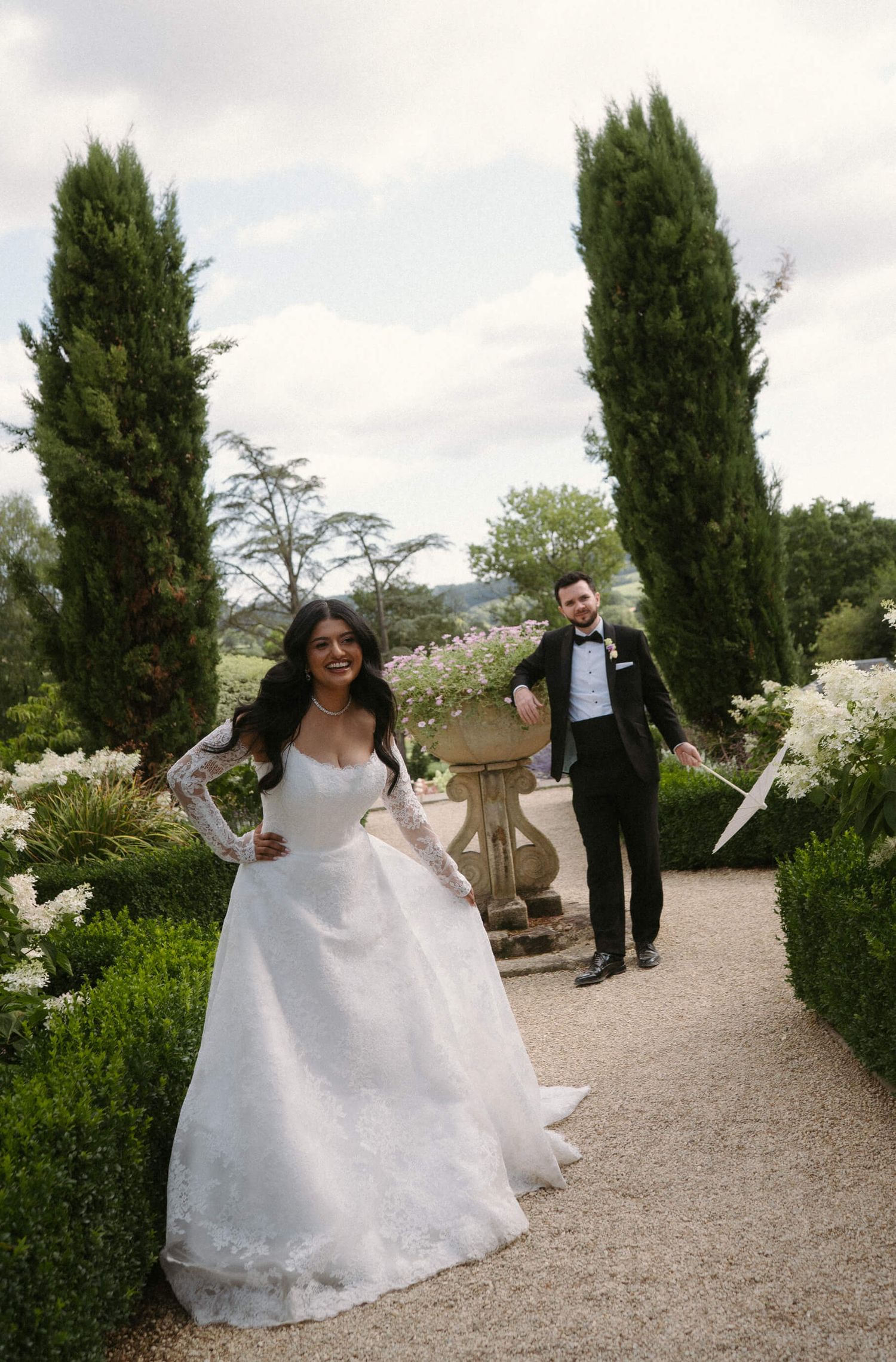 A joyful wedding couple poses in a garden path lined with cypress trees and white flowers.