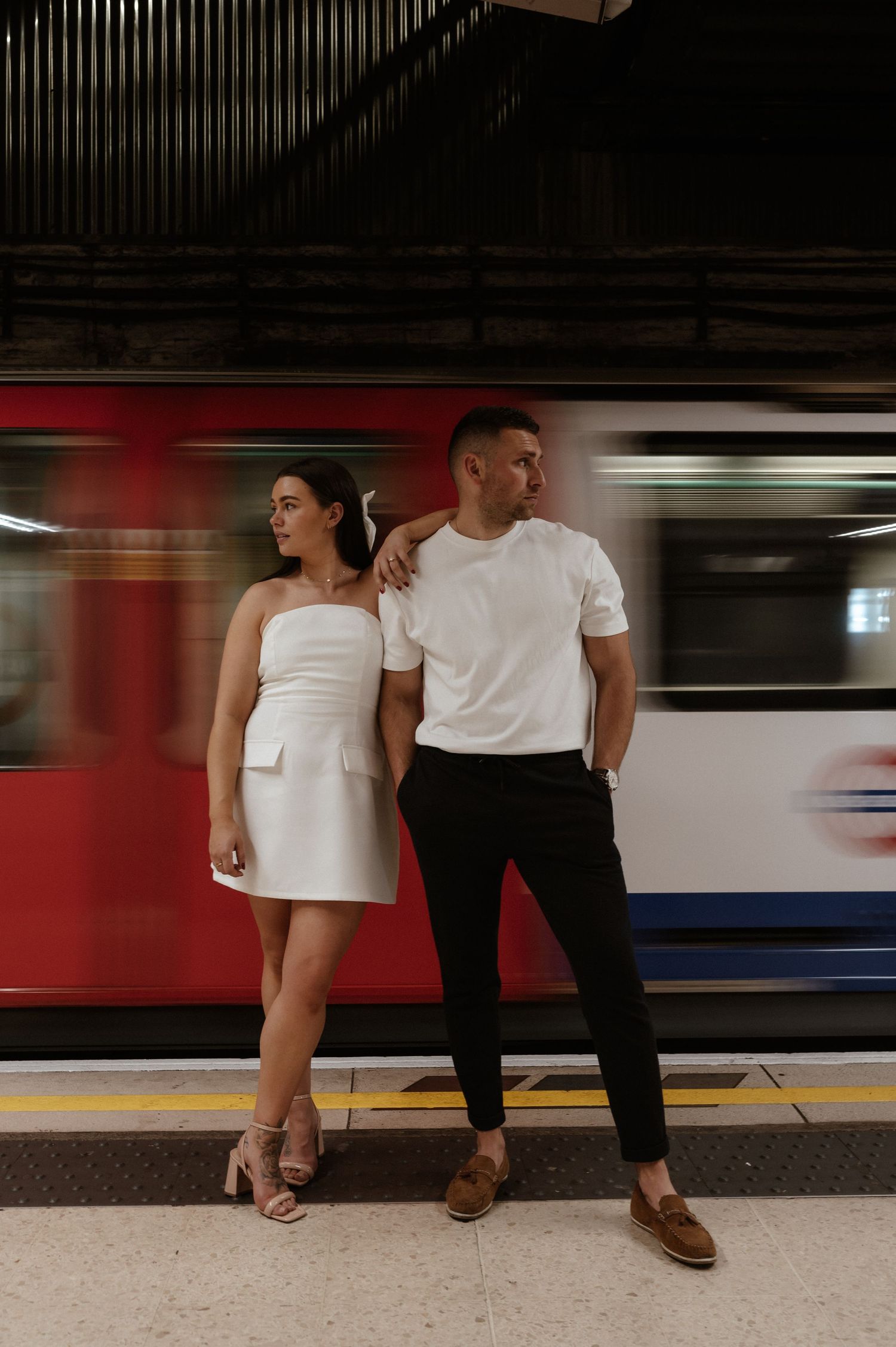 Stylish couple poses against moving subway train in urban station.