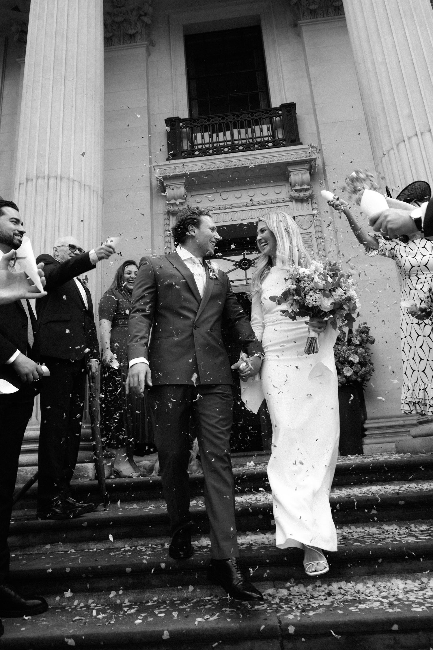 Bride and groom smile and hold hands while walking down stone steps as guests throw confetti outside a grand columned building.