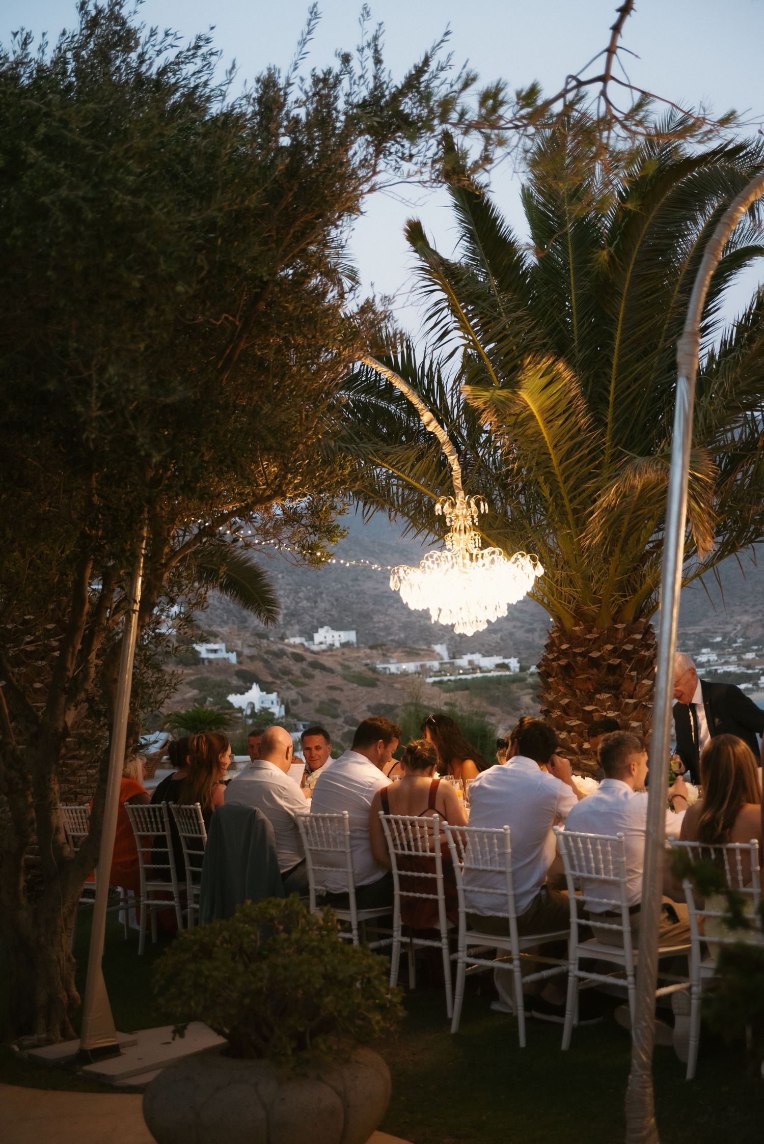 Guests dine at a long outdoor table at dusk beneath a glowing chandelier, palm tree and hillside village views.