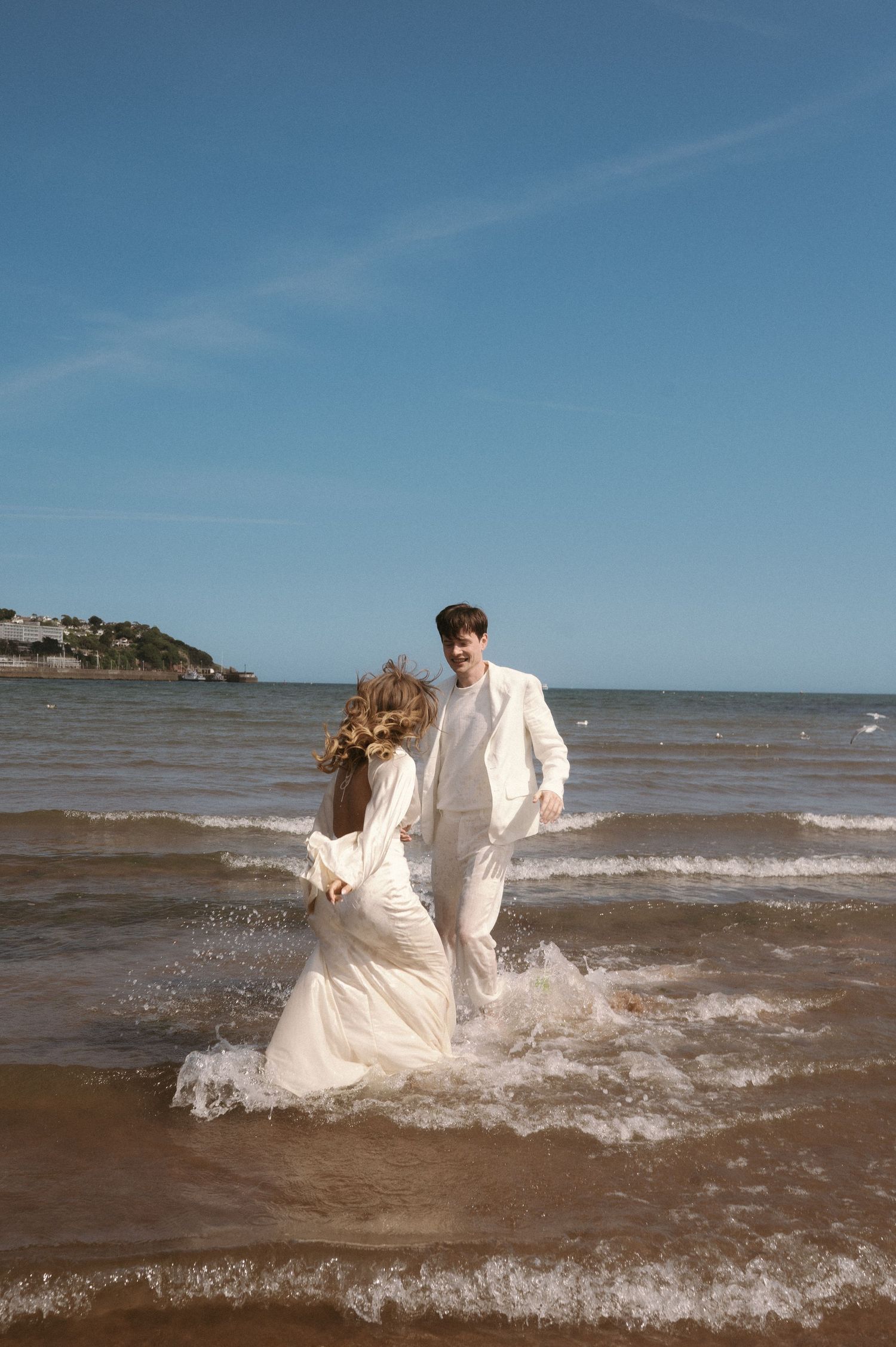 A couple in flowing white attire splashing through ocean waves along a scenic beach.