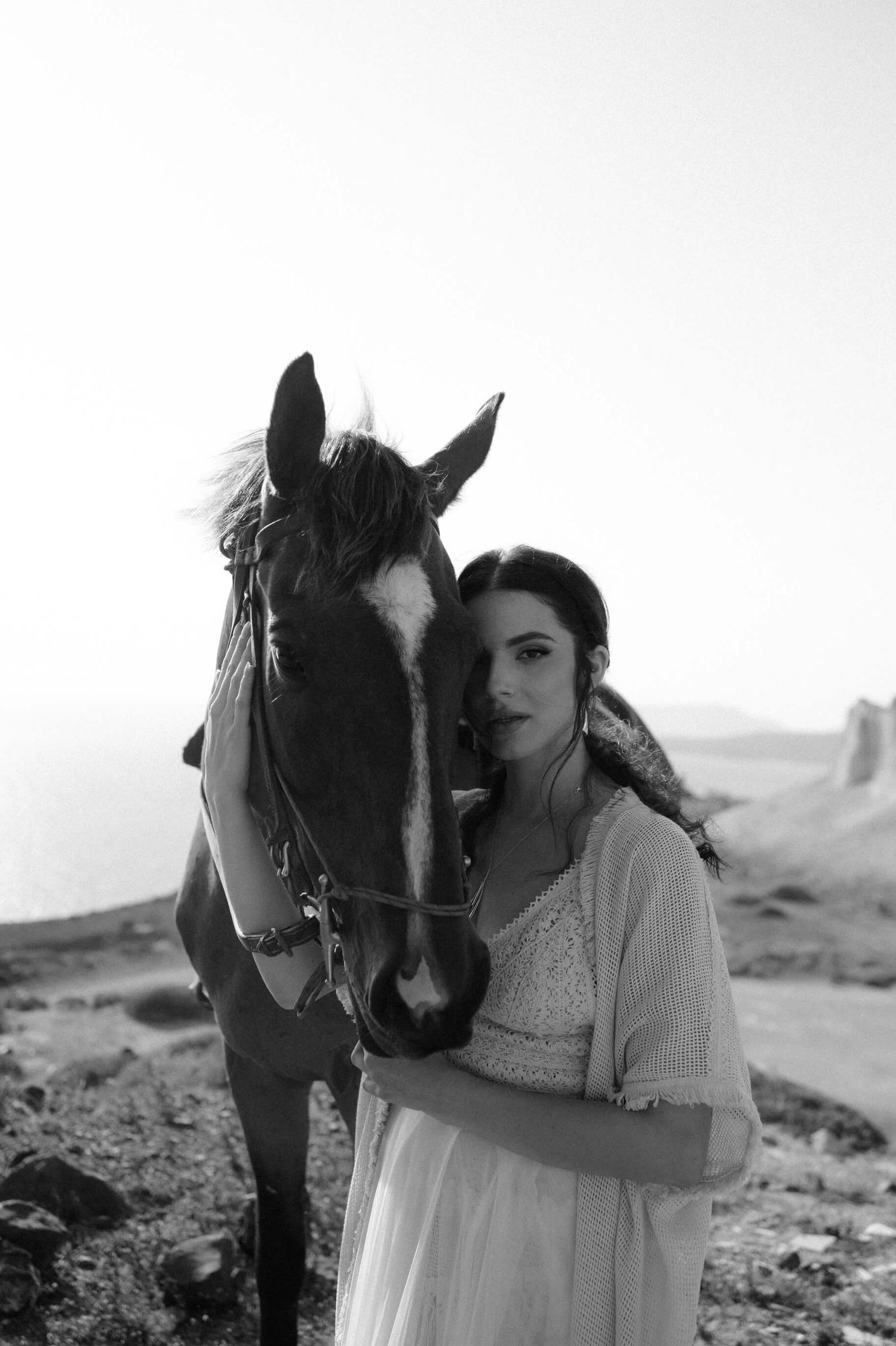 Black and white portrait of a horse and person in a white dress against a desert landscape.